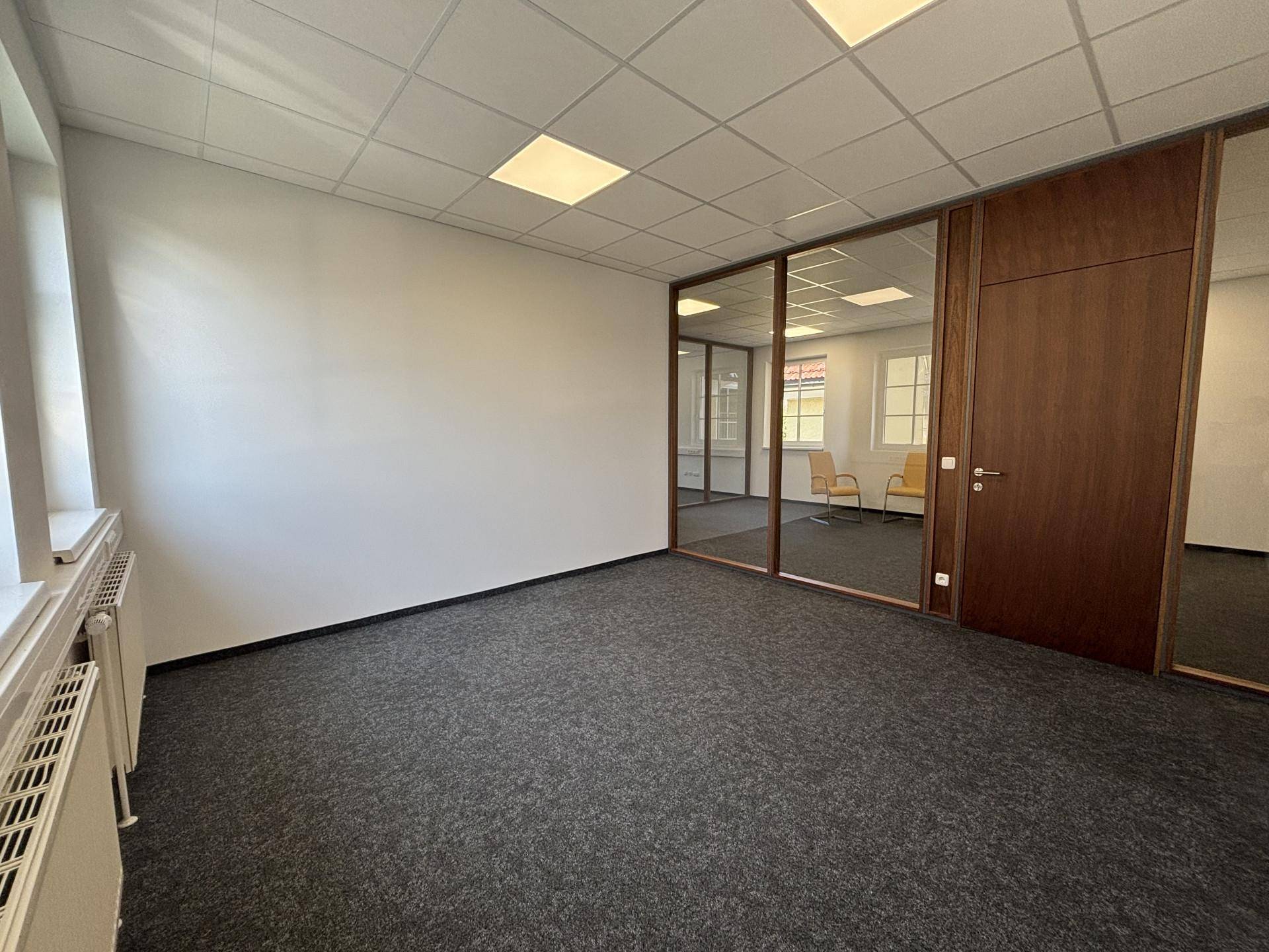 Helles Büro mit grauem Teppichboden, weißen Wänden und einer modernen Glasabtrennung zu einem weiteren Raum.