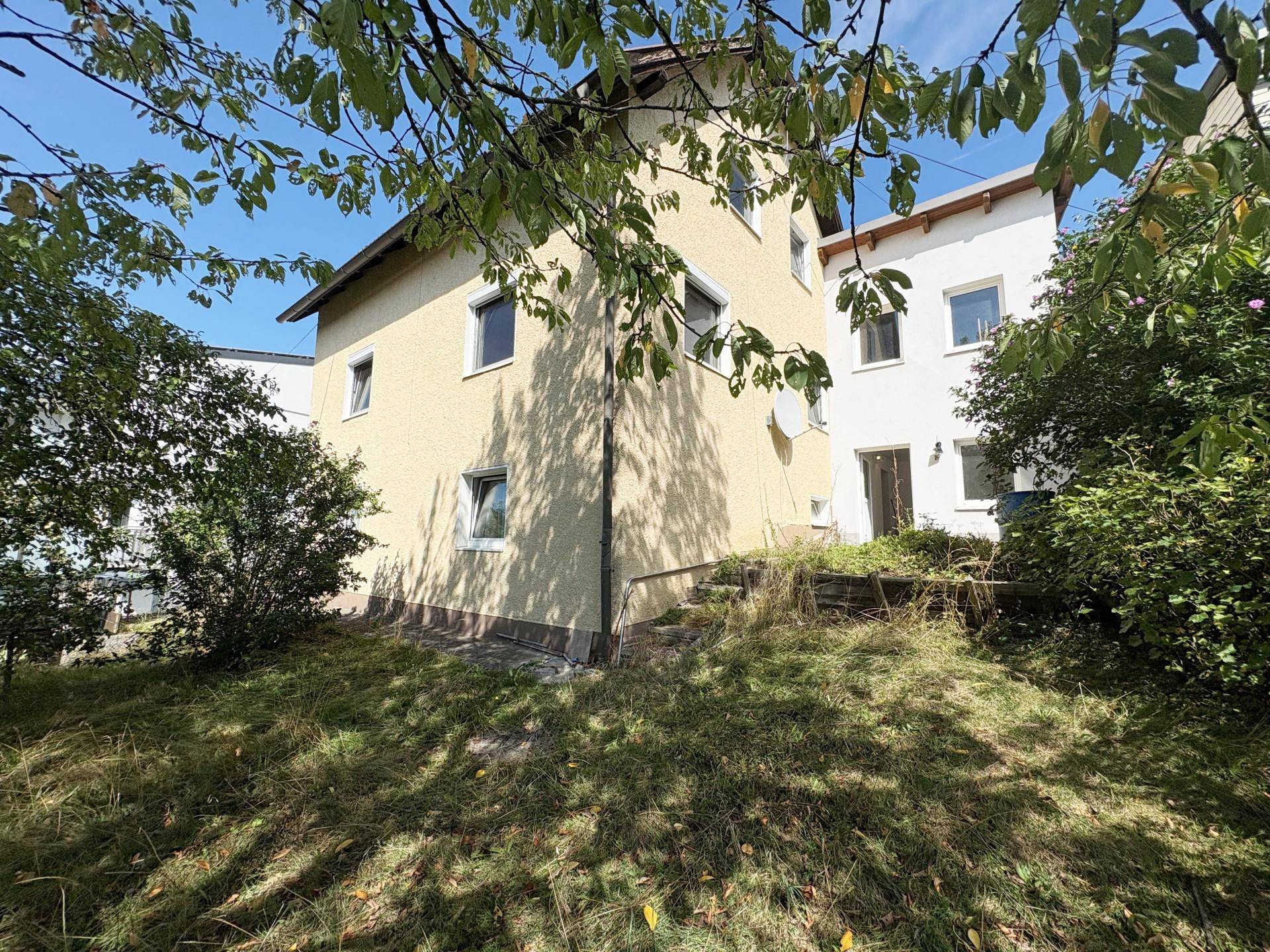 Seitenansicht des Hauses mit bewachsenem Gartenbereich und Bäumen im Vordergrund.