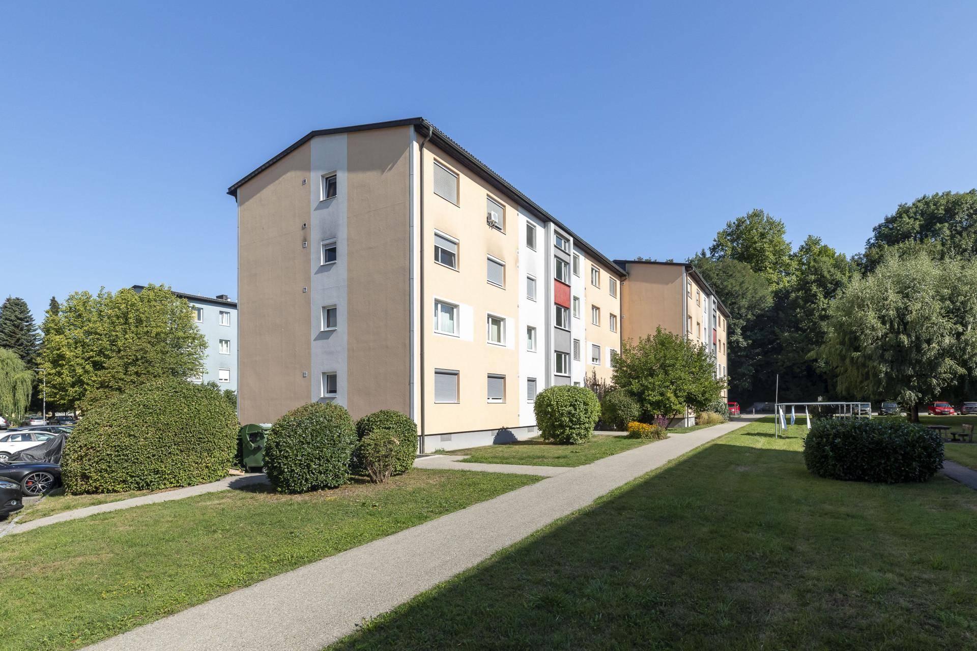 Mehrfamilienhaus mit gepflegter Grünanlage und Parkplätzen unter blauem Himmel.