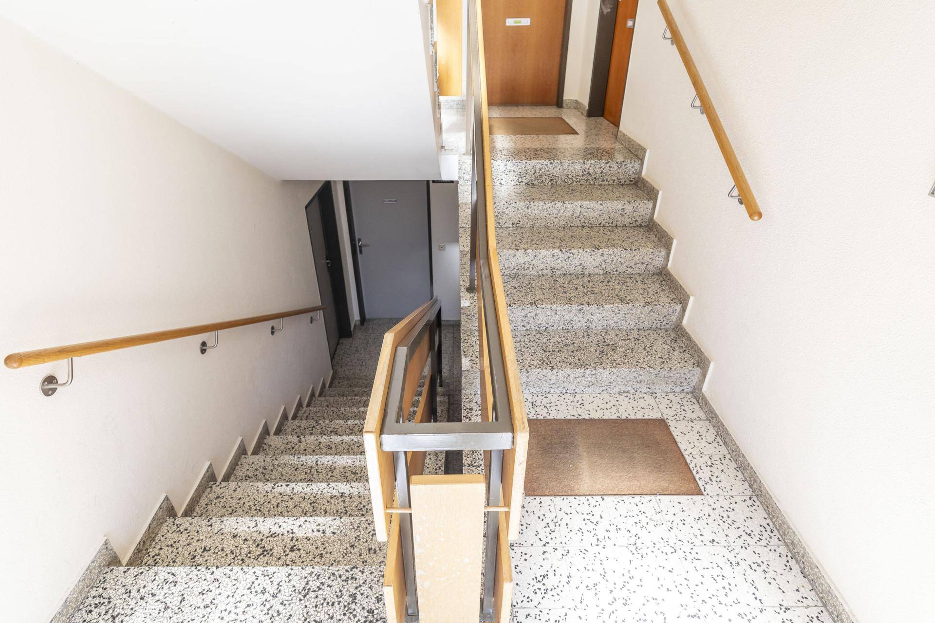 Treppenhaus mit Terrazzoboden, Holzgeländer und natürlichem Lichteinfall.