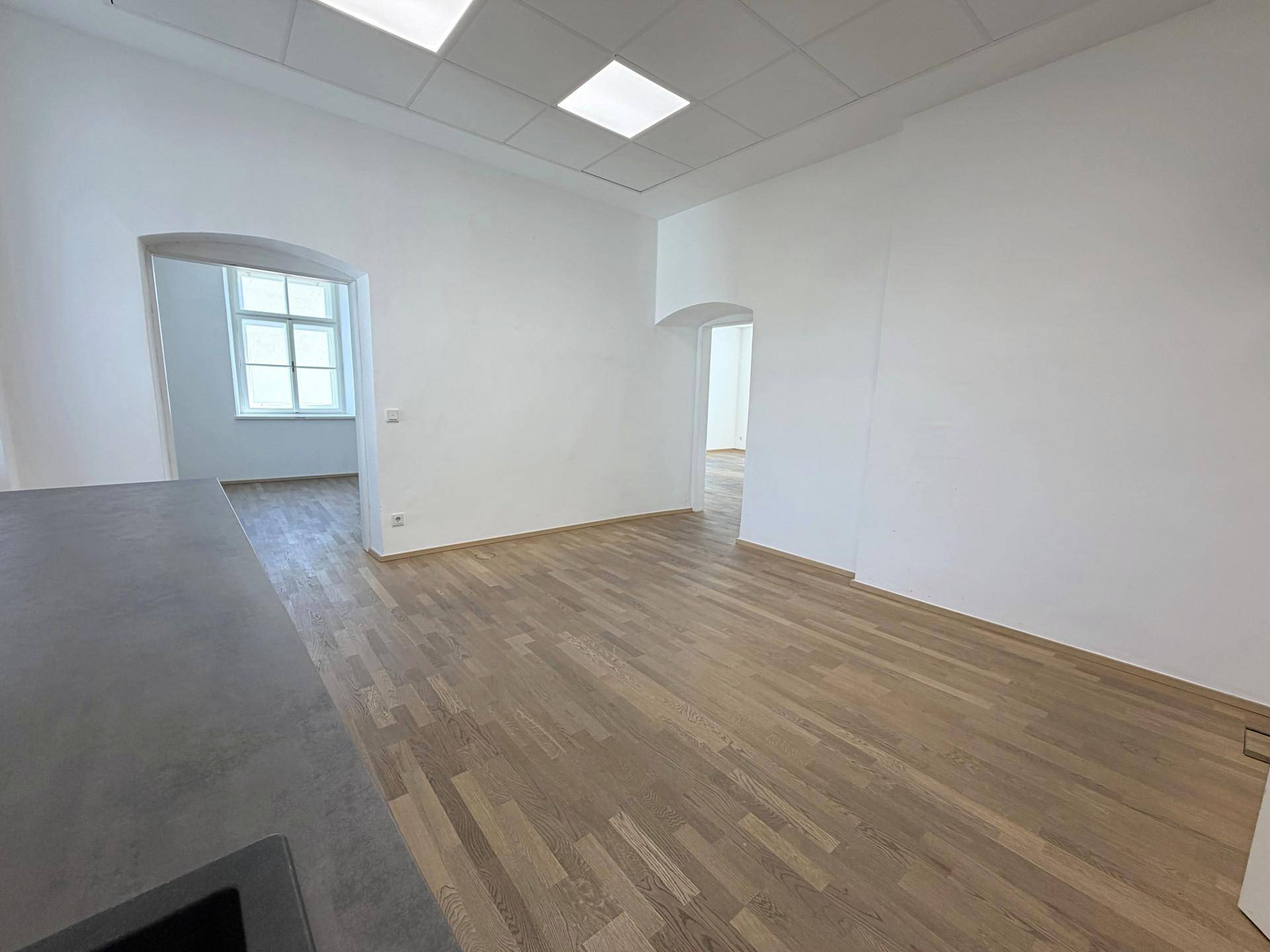 Geräumiger Raum mit Holzboden und weißen Wänden, der durch offene Bögen weitere Bereiche verbindet.