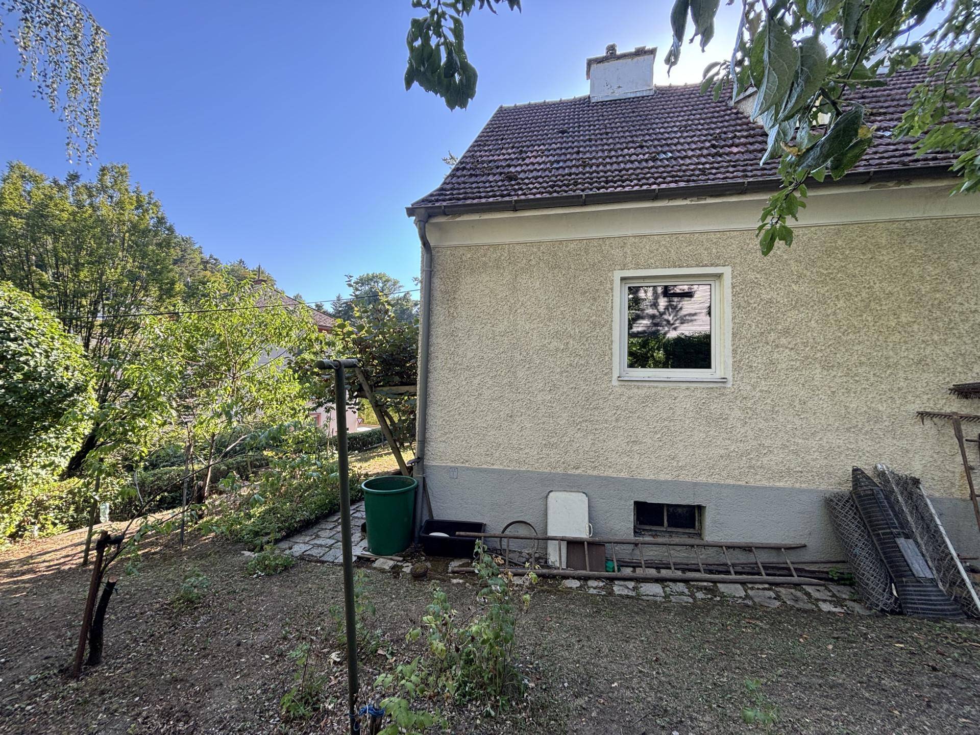 Seitenansicht des Hauses mit einfacher Putzfassade und einem unaufgeräumten Gartenbereich.