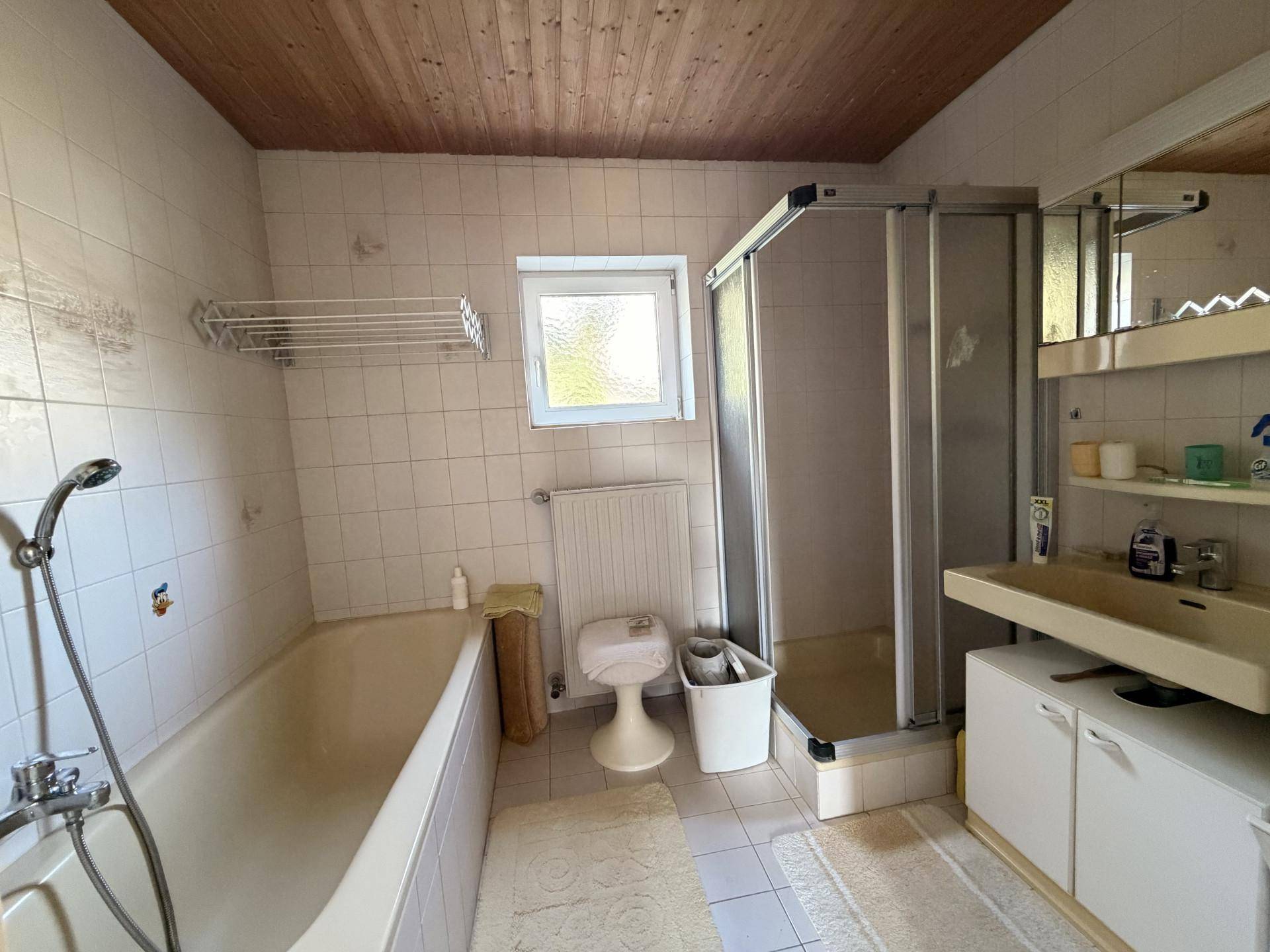 Badezimmer mit Badewanne, Duschkabine, Waschbecken und Fenster, ausgestattet mit weißen Fliesen.