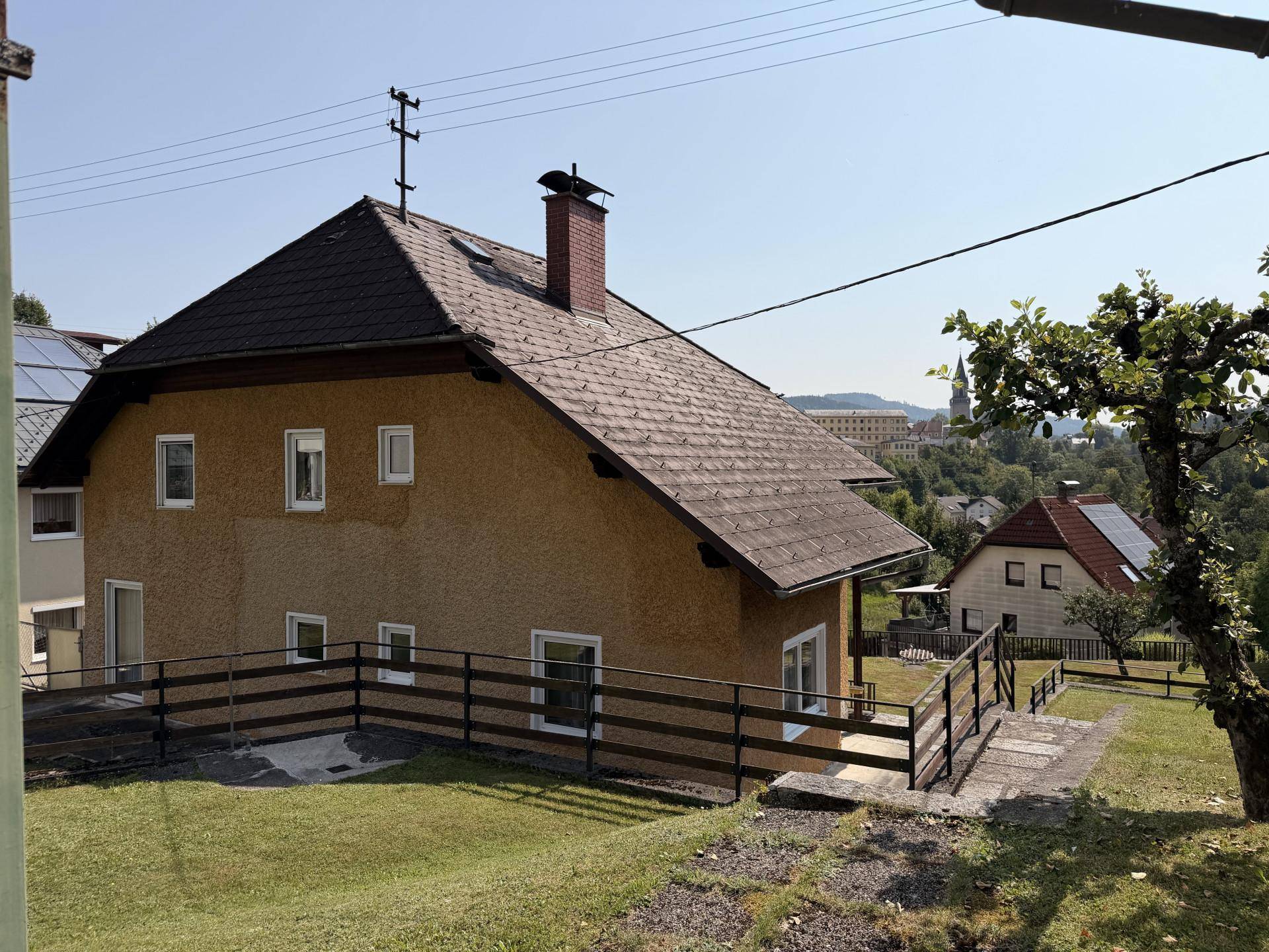 Ansicht des Hauses mit gelber Fassade und braunem Dach, umgeben von grüner Landschaft und Fernblick.