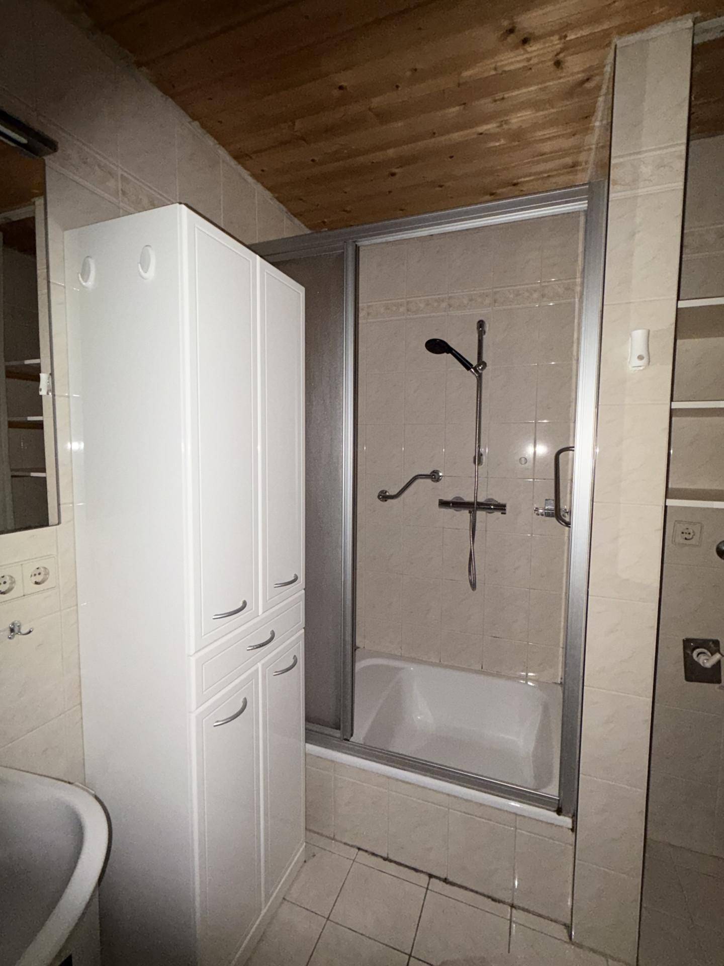 Badezimmer mit Duschwanne, weißen Fliesen und einem praktischen Hochschrank, unter einer Holzdecke.
