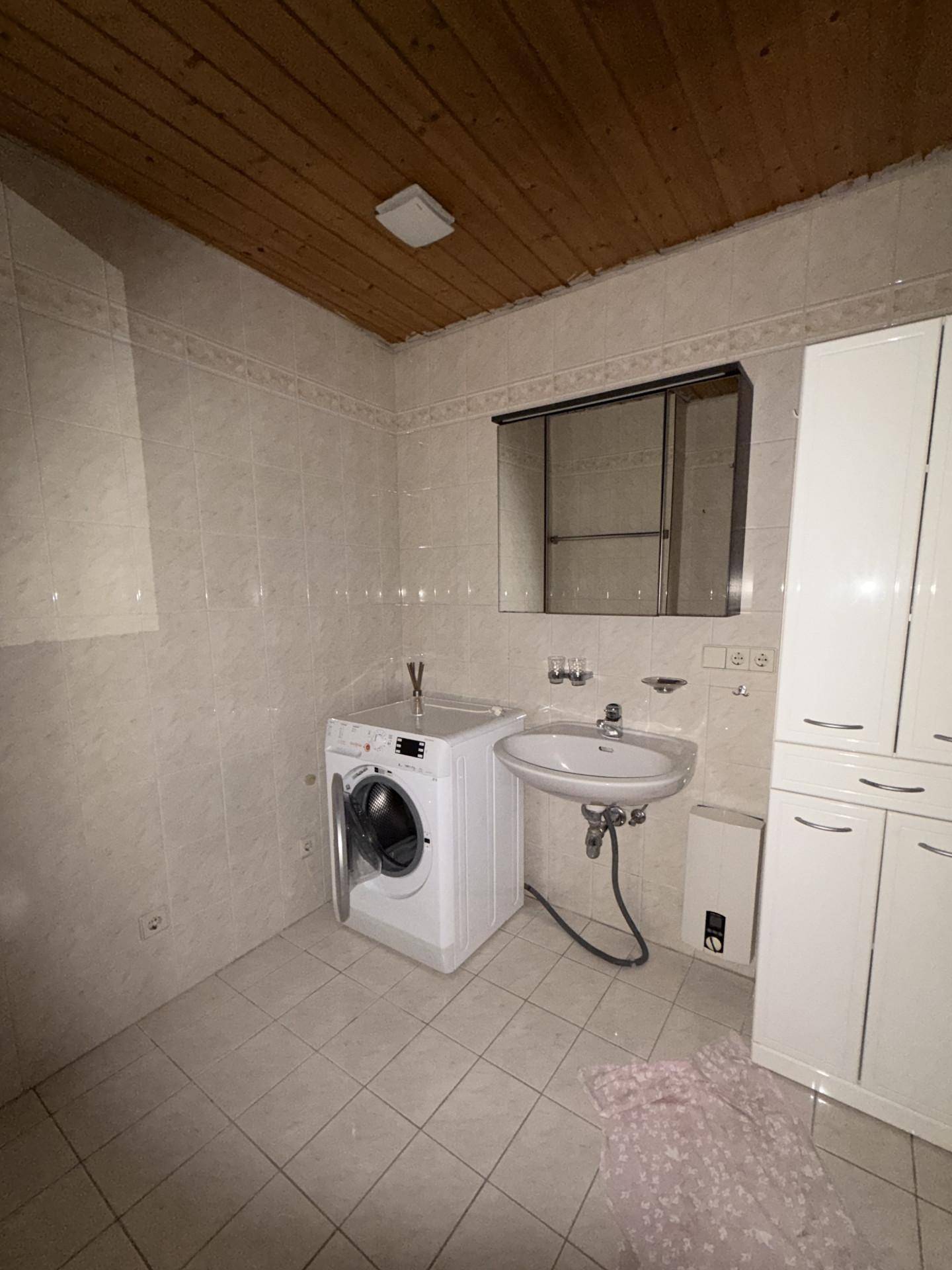 Badezimmer mit Waschmaschine, Waschbecken und weiß gefliesten Wänden, ergänzt durch eine Holzdecke.