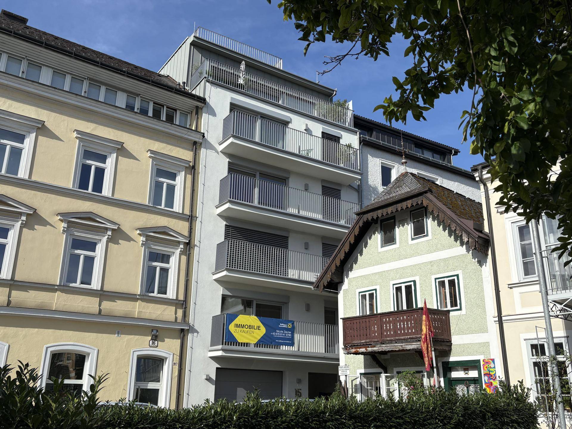 Ansicht einer Straße mit einer Mischung aus modernen und traditionellen Gebäuden, darunter ein Neubau mit Balkonen und Dachterrasse.