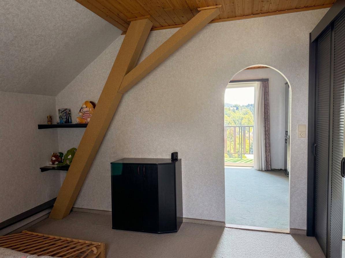 Dachgeschossraum mit schräger Decke, sichtbarem Holzbalken und einem Bogenzugang zu einem weiteren Zimmer.