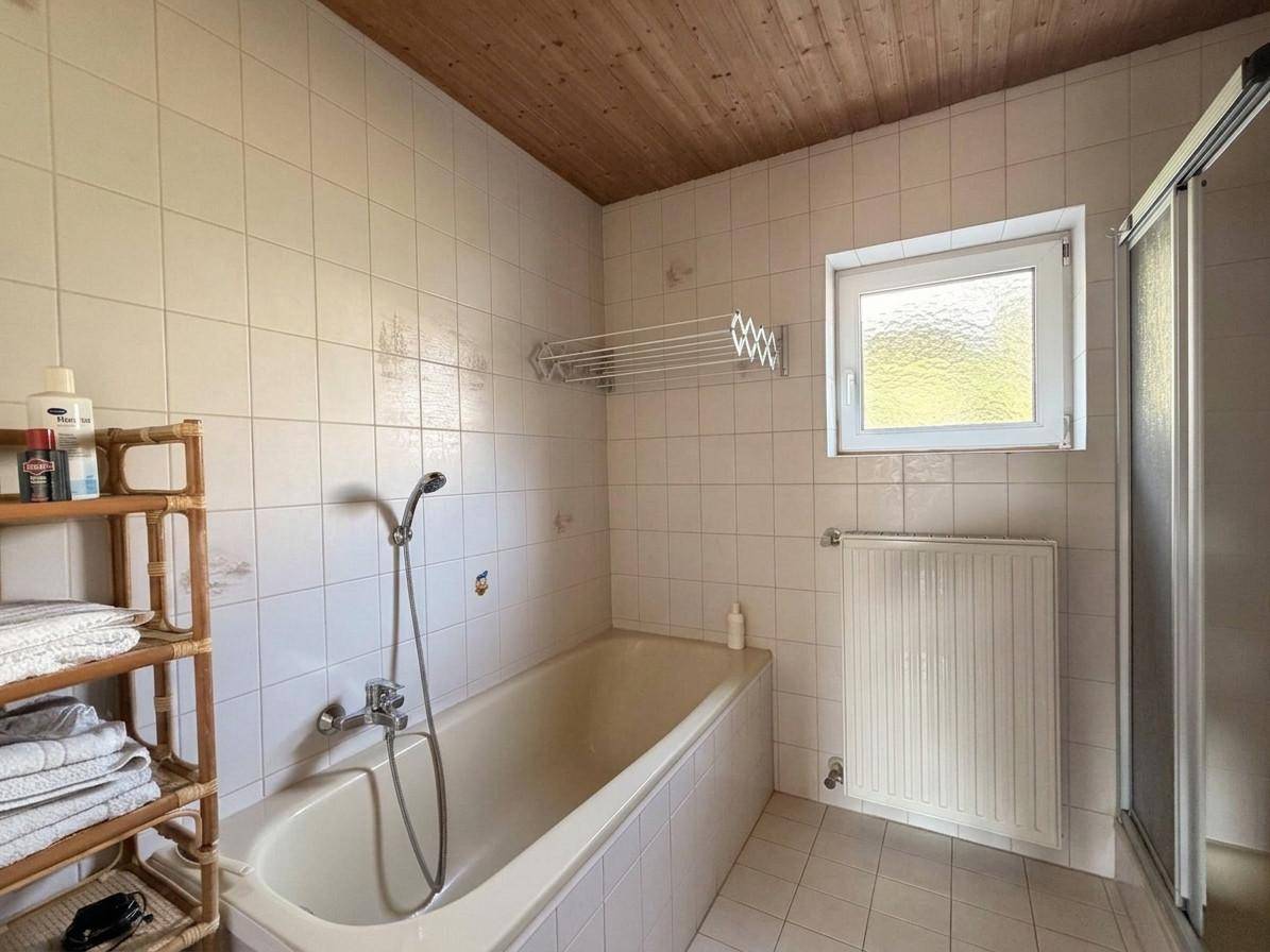 Helles Badezimmer mit Badewanne, Dusche, WC und einem Fenster, alles in weißen Fliesen gehalten.
