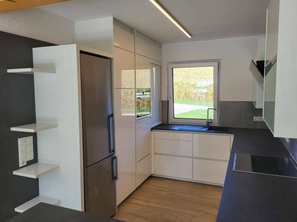 Moderne Küche mit weißen Hochglanzfronten, dunkler Arbeitsplatte und Fenster mit Blick ins Grüne.