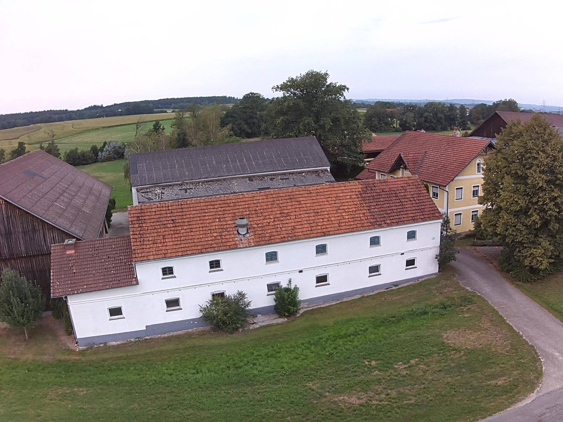 Ländliches Nebengebäude mit rotem Ziegeldach und weißer Fassade, eingebettet in grüne Landschaft.
