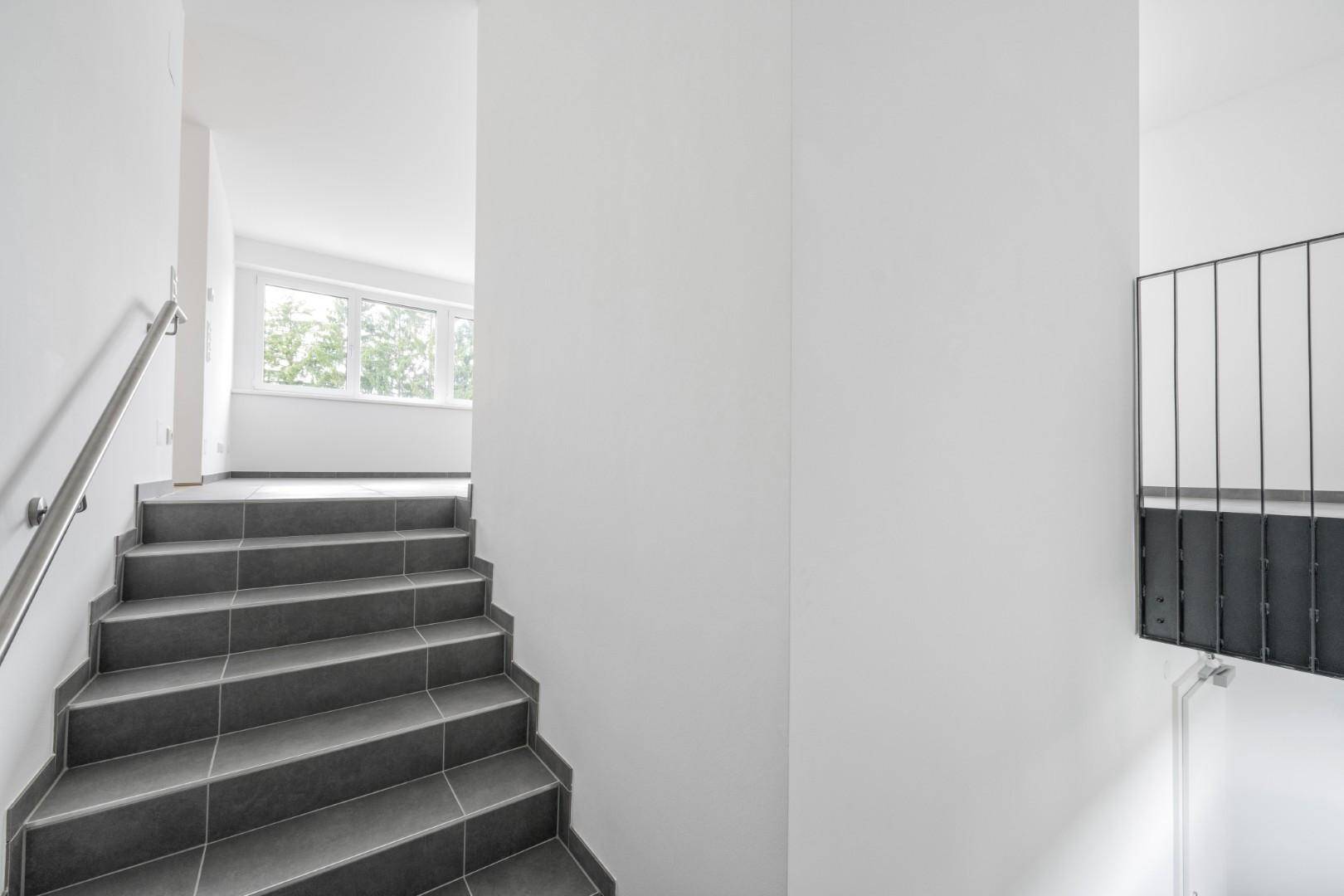 Modernes Treppenhaus mit grauen Fliesen und schlichtem Metallgeländer, führt zu den oberen Etagen.