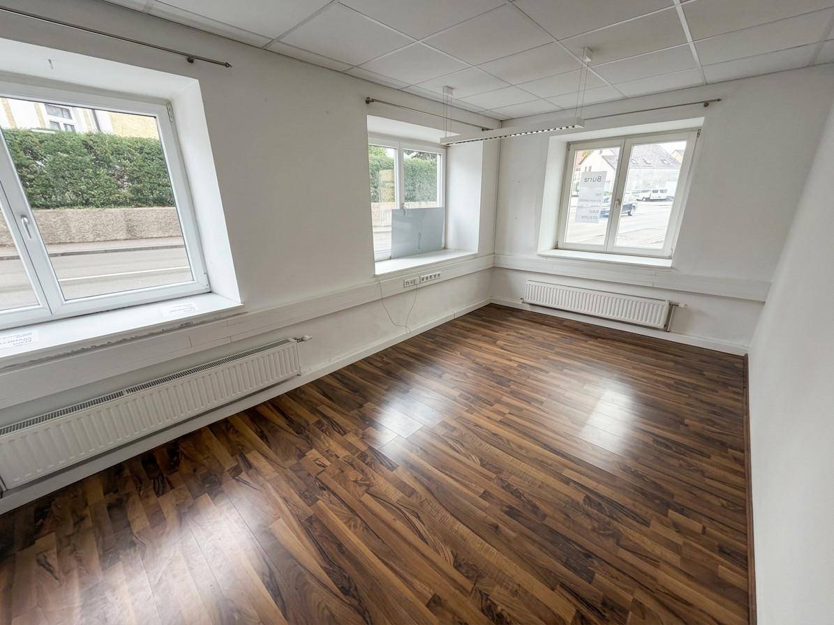 Leerer Büroraum mit dunklem Laminatboden, weißen Wänden und mehreren Fenstern, die viel Tageslicht hereinlassen.