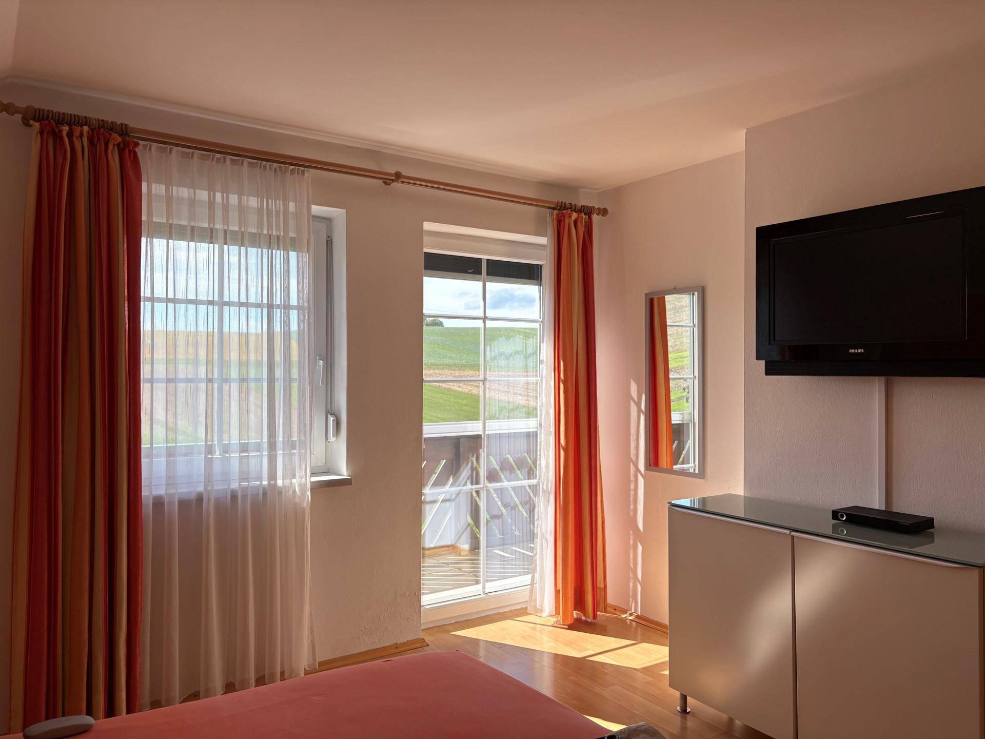 Schlafzimmer mit Holzboden, TV und Zugang zu einem Balkon mit weitem Blick über die Felder.