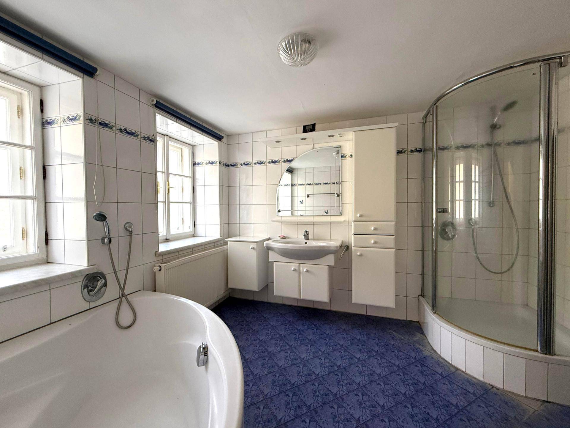 Geräumiges Badezimmer mit blauen Fliesen, Badewanne, Dusche und zwei Fenstern.
