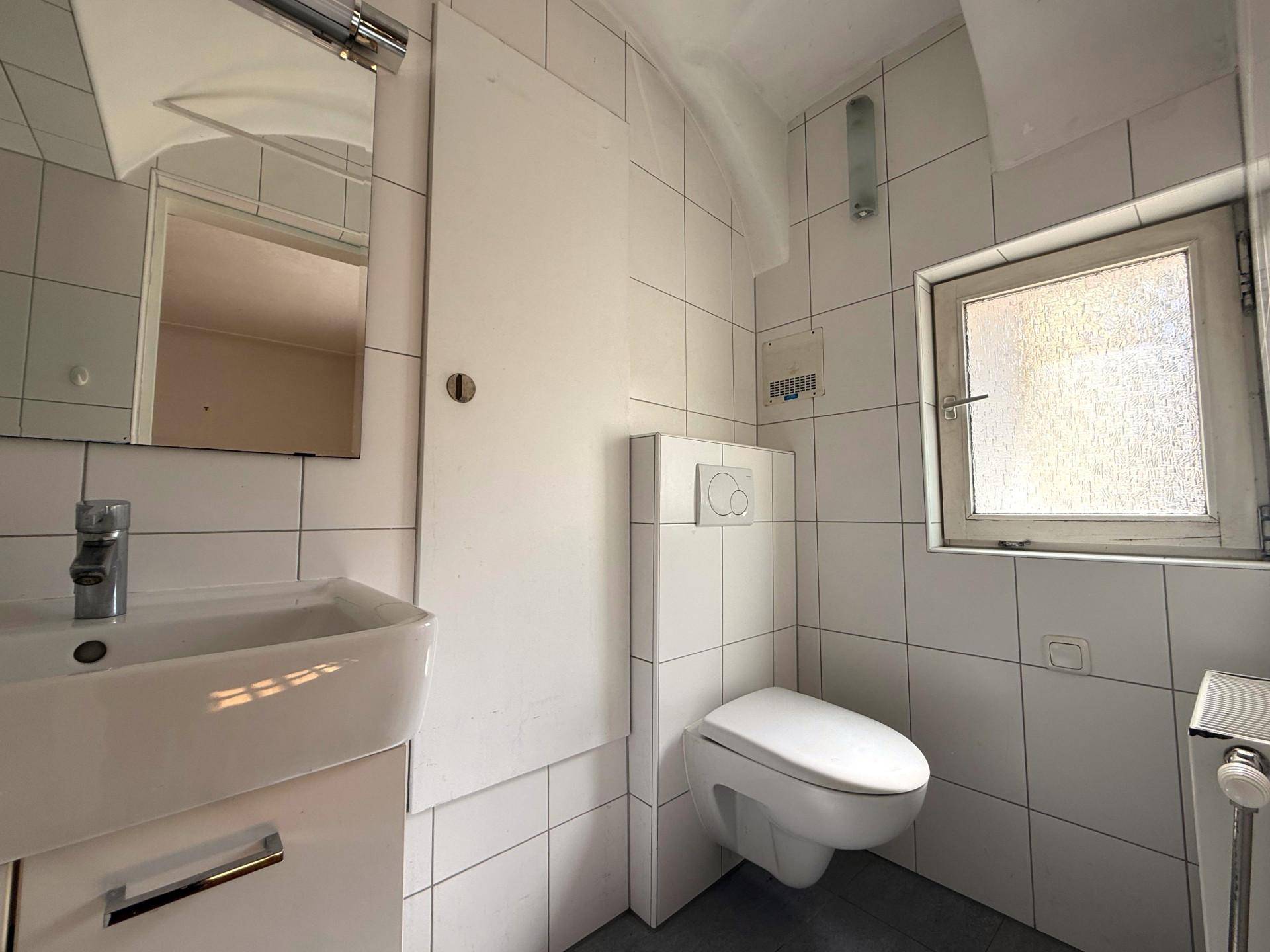 Helles WC mit Wand-Toilette, Waschbecken und Fenster, komplett gefliest.
