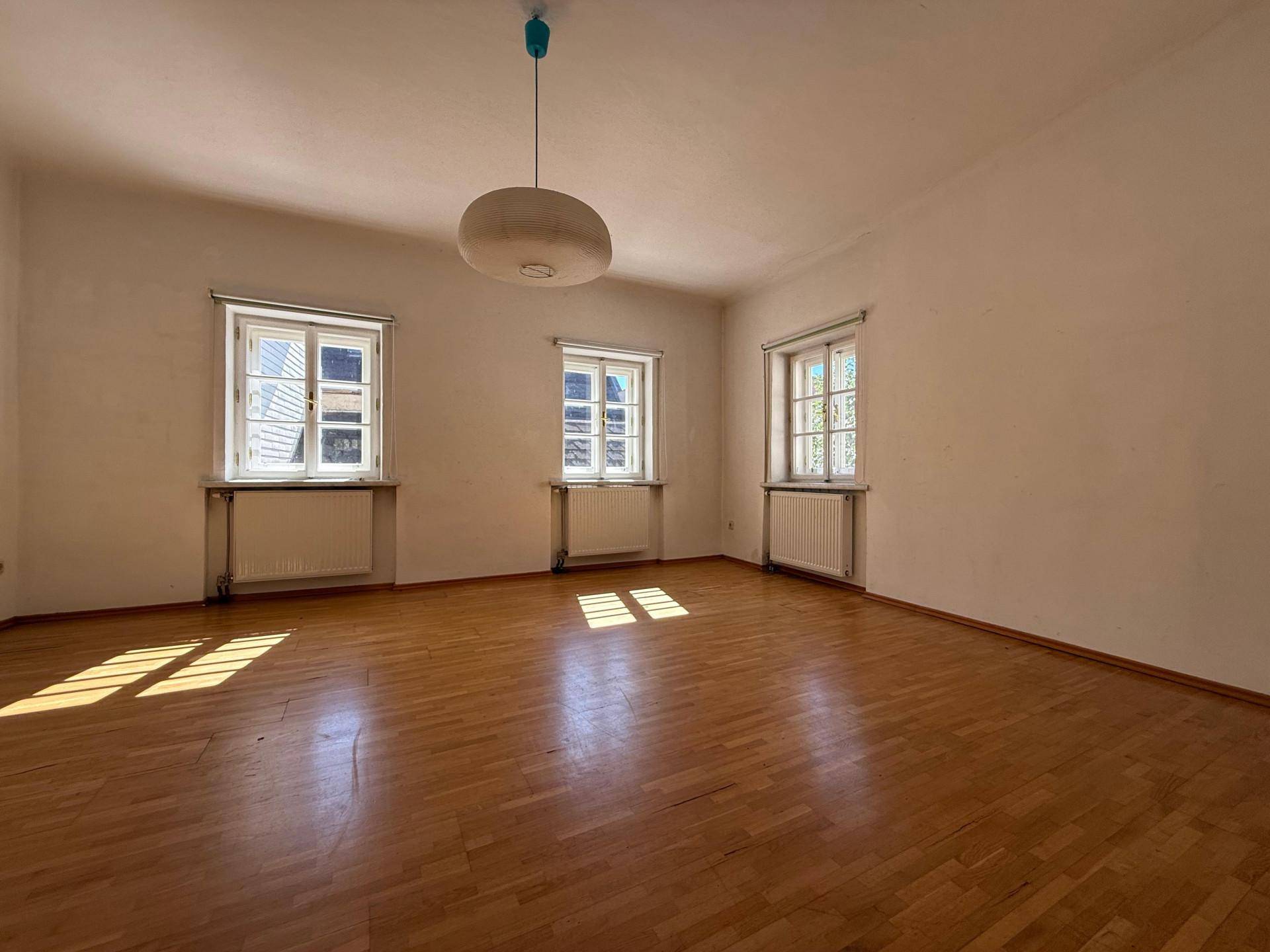Helles Wohnzimmer mit Holzboden und drei Fenstern, die viel Tageslicht hereinlassen.