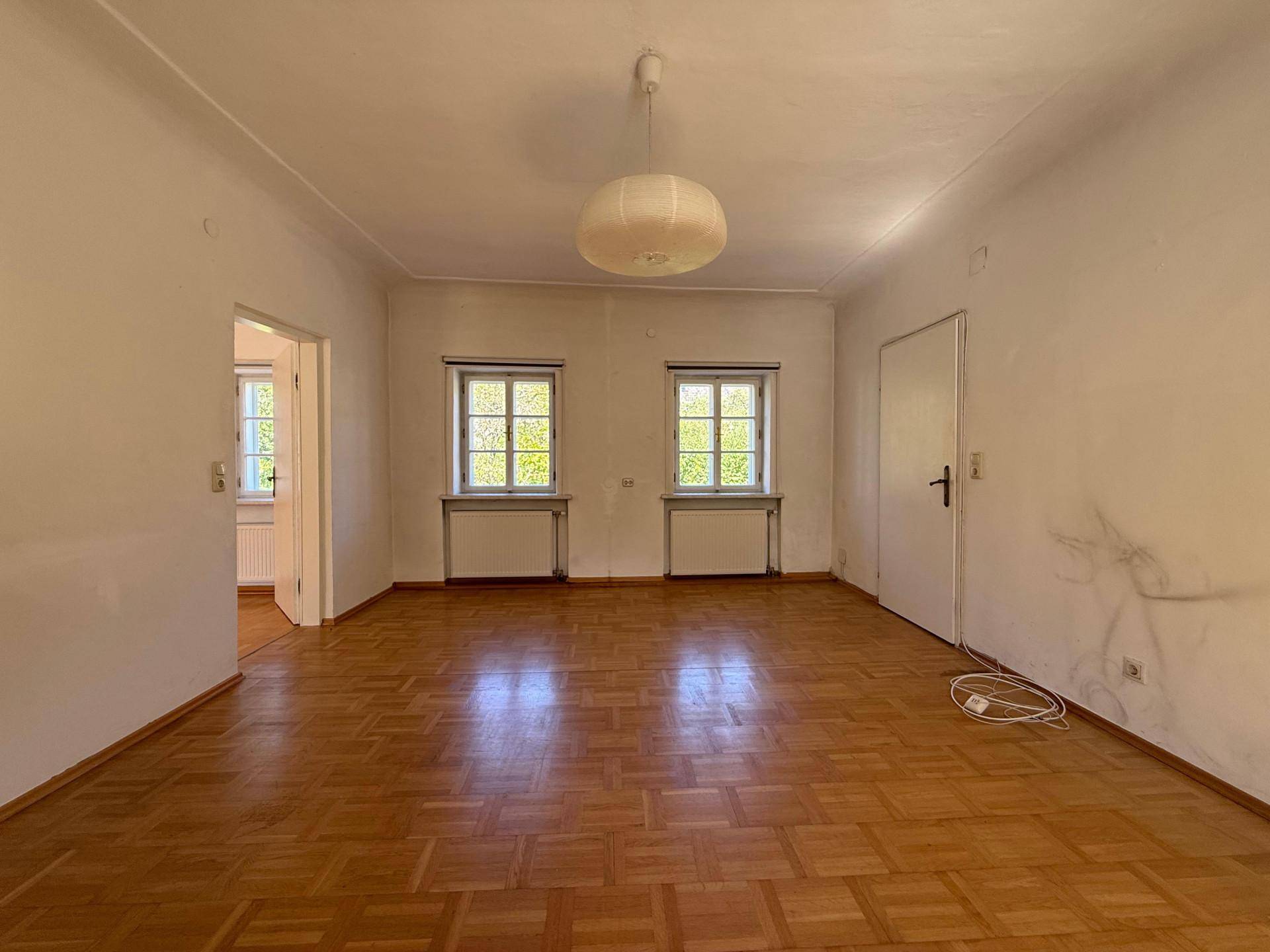 Geräumiges Wohnzimmer mit Holzboden, zwei Fenstern und einer modernen Deckenleuchte.