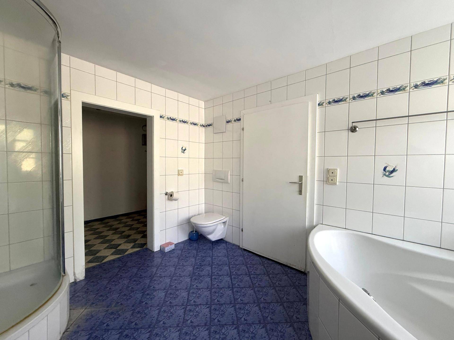 Badezimmer mit blauen Bodenfliesen, Wand-WC, Dusche und Zugang zu einem weiteren Raum.