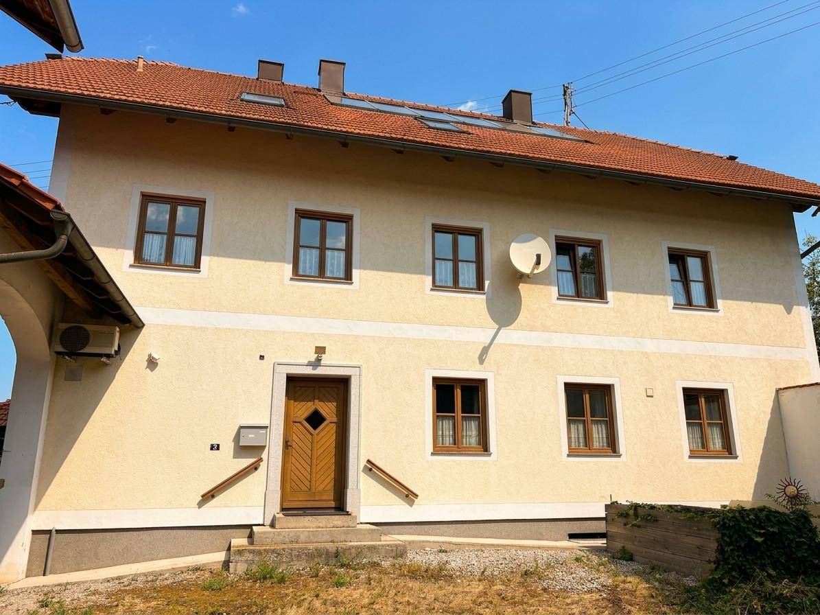 Vorderansicht des Hauses mit heller Fassade, braunen Fenstern und Satellitenschüssel am Giebel.