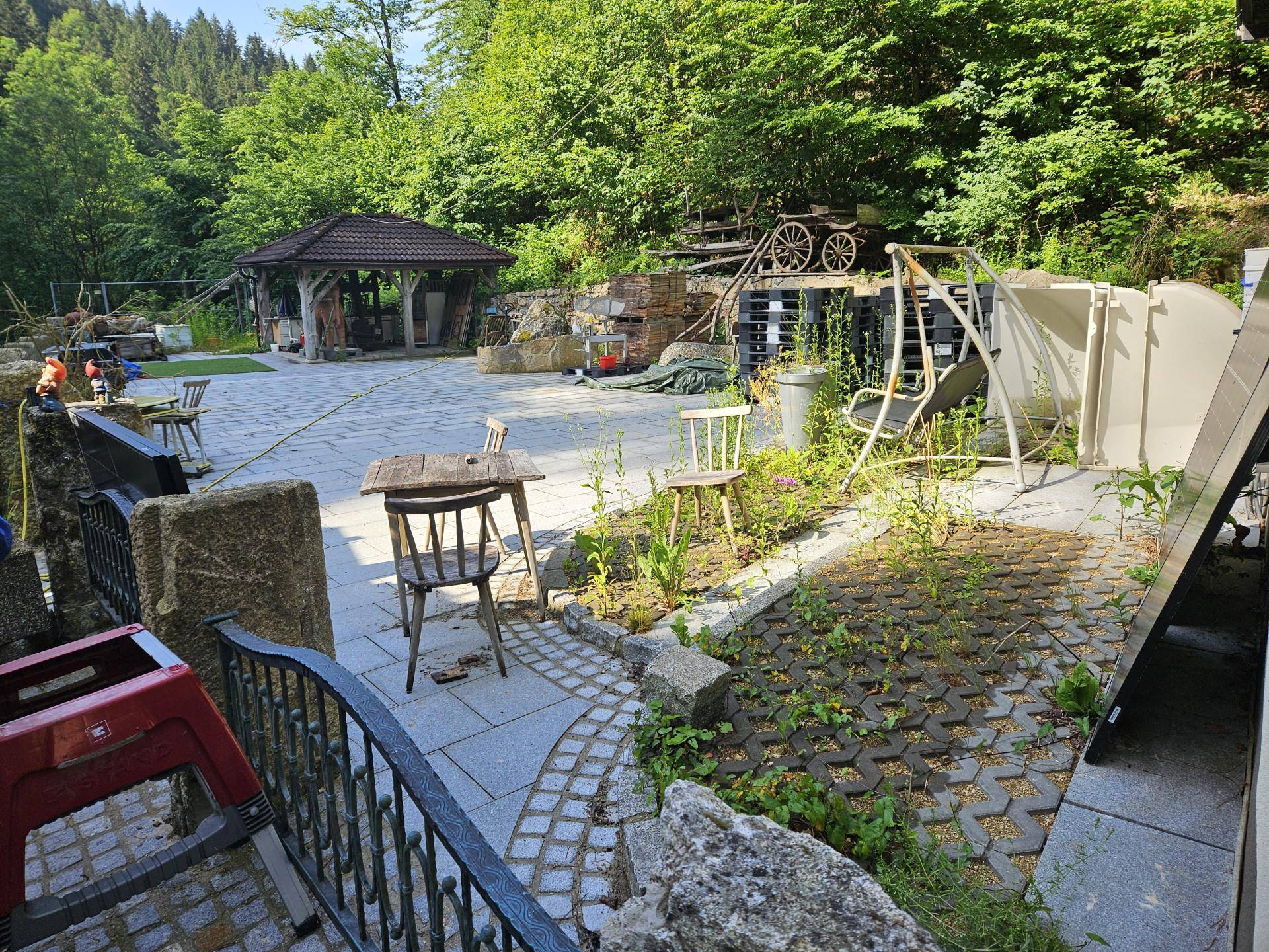 Weitläufige gepflasterte Terrasse mit Sitzgelegenheiten und einem kleinen Gartenbereich, umgeben von Wald.