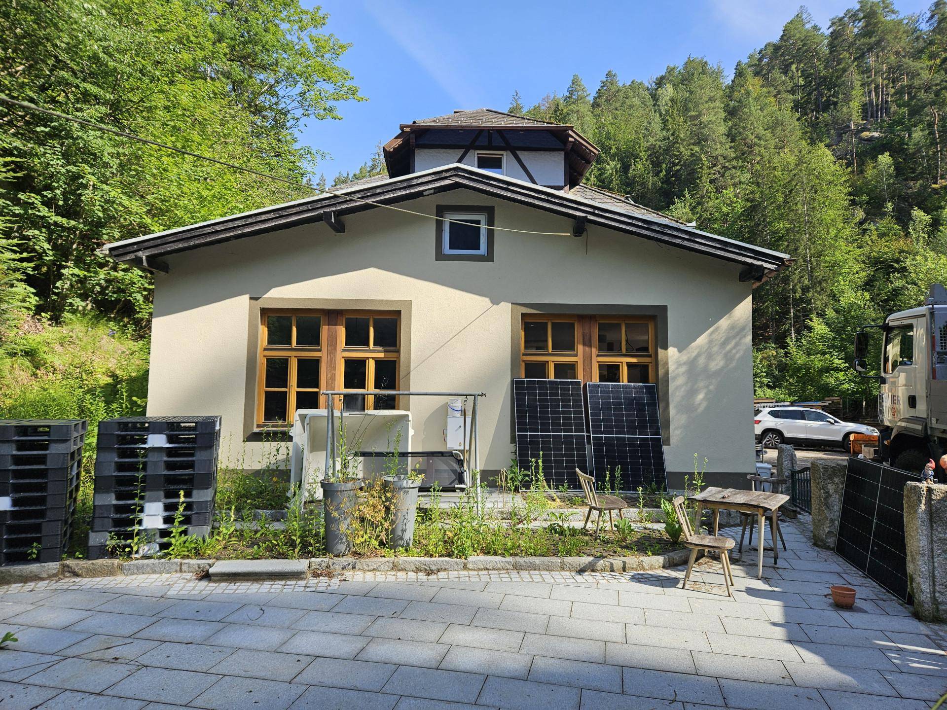 Kleineres Gebäude mit Holzfenstern und Solarmodulen an der Fassade, umgeben von Bäumen.