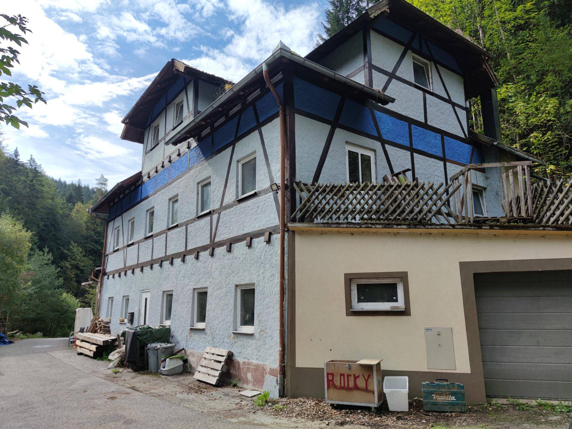 Seitenansicht eines älteren Gebäudes mit Fachwerkdetails und einer Garage, umgeben von viel Grün.