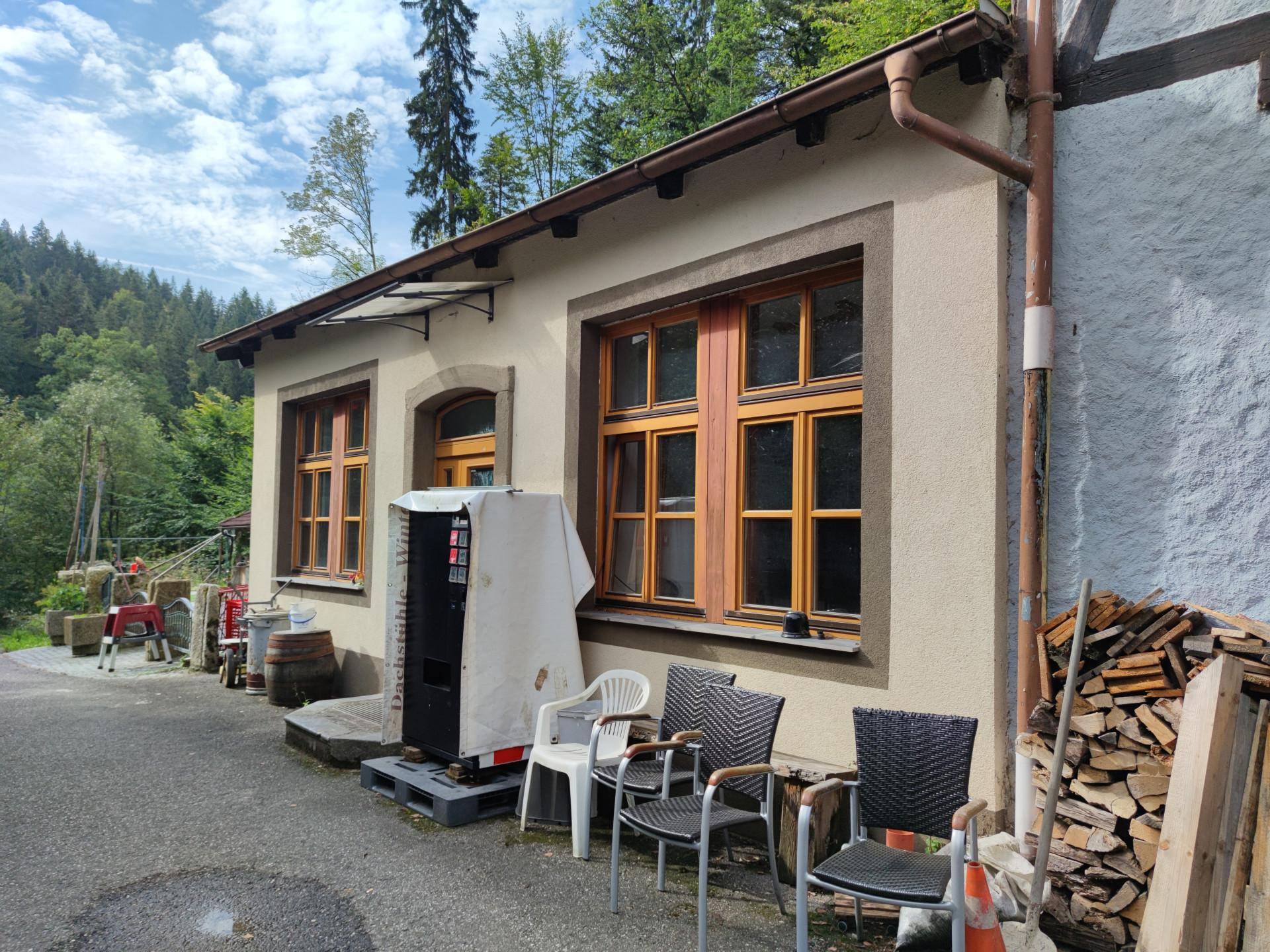 Fassade eines Gebäudes mit Holztüren und Fenstern, davor stehen Stühle und ein Verkaufsautomat.