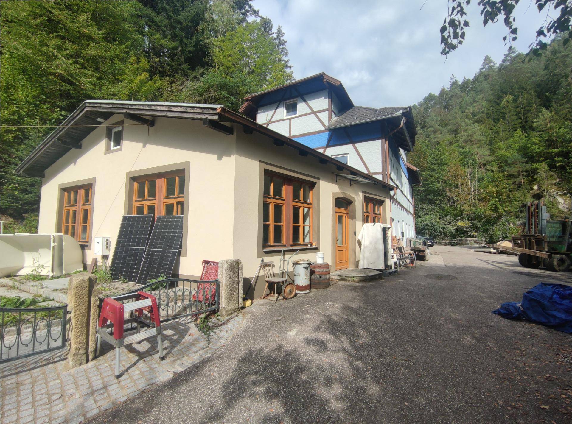 Ansicht eines Gebäudekomplexes mit verschiedenen Baustilen und Solarmodulen, umgeben von üppigem Grün.