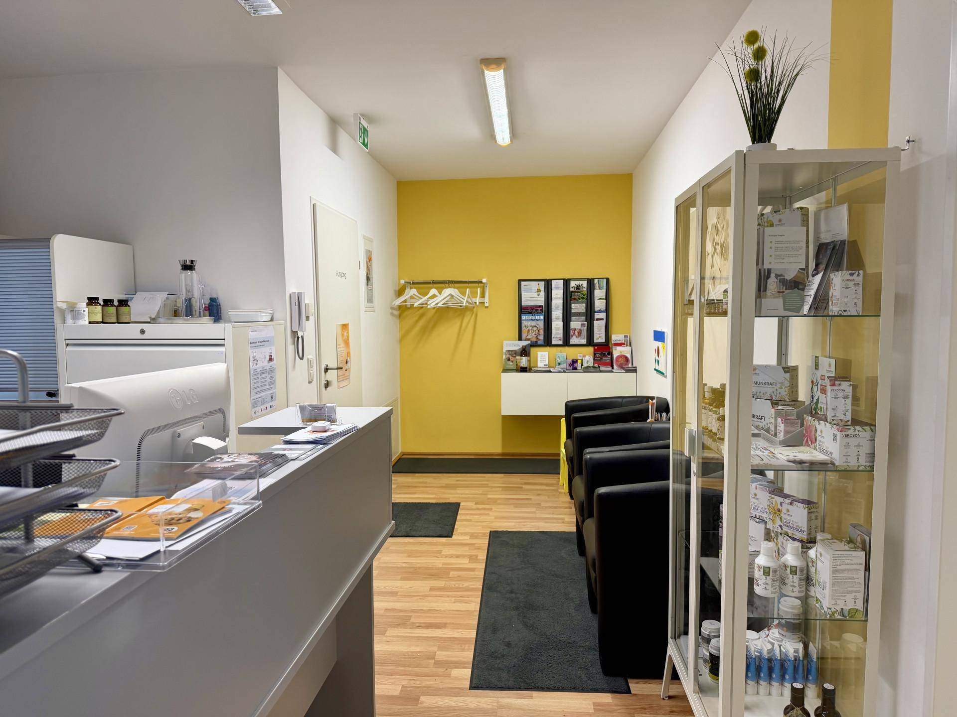 Heller Empfangsbereich mit Rezeption, Wartebereich und einem Verkaufsregal an einer gelben Wand.