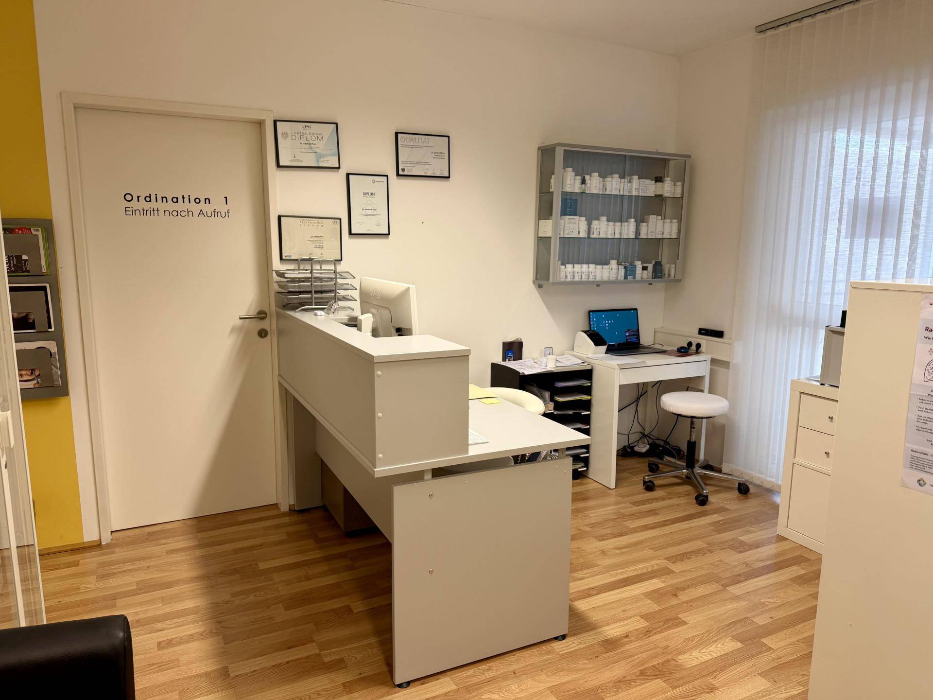 Büro- oder Behandlungsraum mit Empfangstheke, Schreibtisch und einem Fenster mit Jalousien.