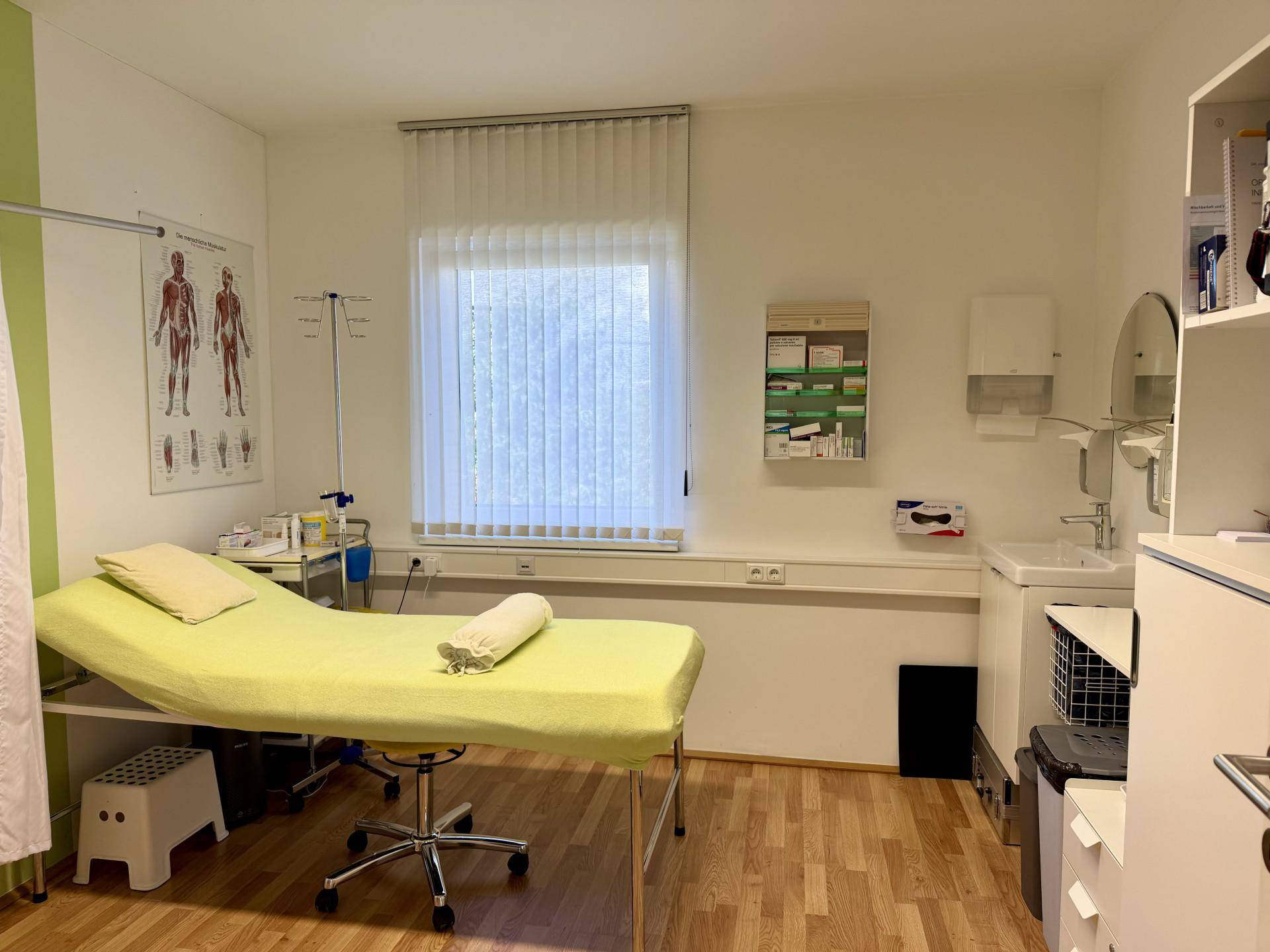 Behandlungsraum mit Untersuchungsliege, Waschbecken und medizinischen Diagrammen an der Wand.