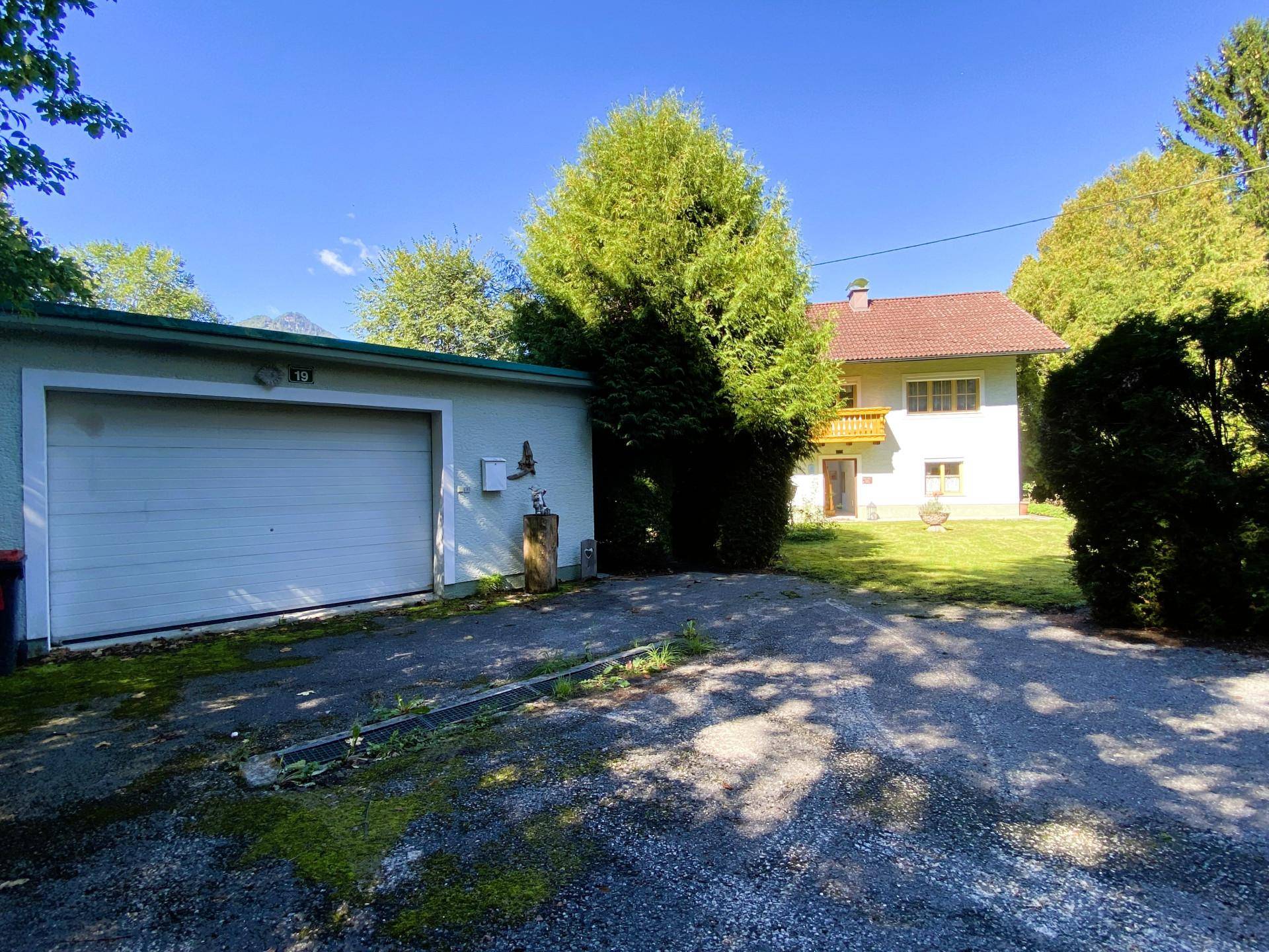 Garage mit Zufahrt und dem Haupthaus im Hintergrund, umgeben von Bäumen.