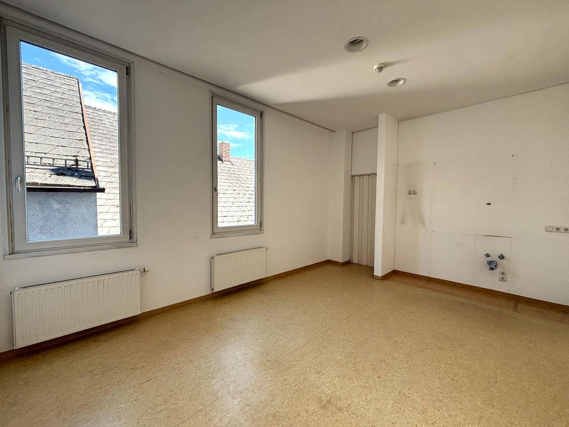 Raum mit Linoleumboden und zwei Fenstern, die einen Blick auf Dächer bieten, mit sichtbaren Anschlüssen.