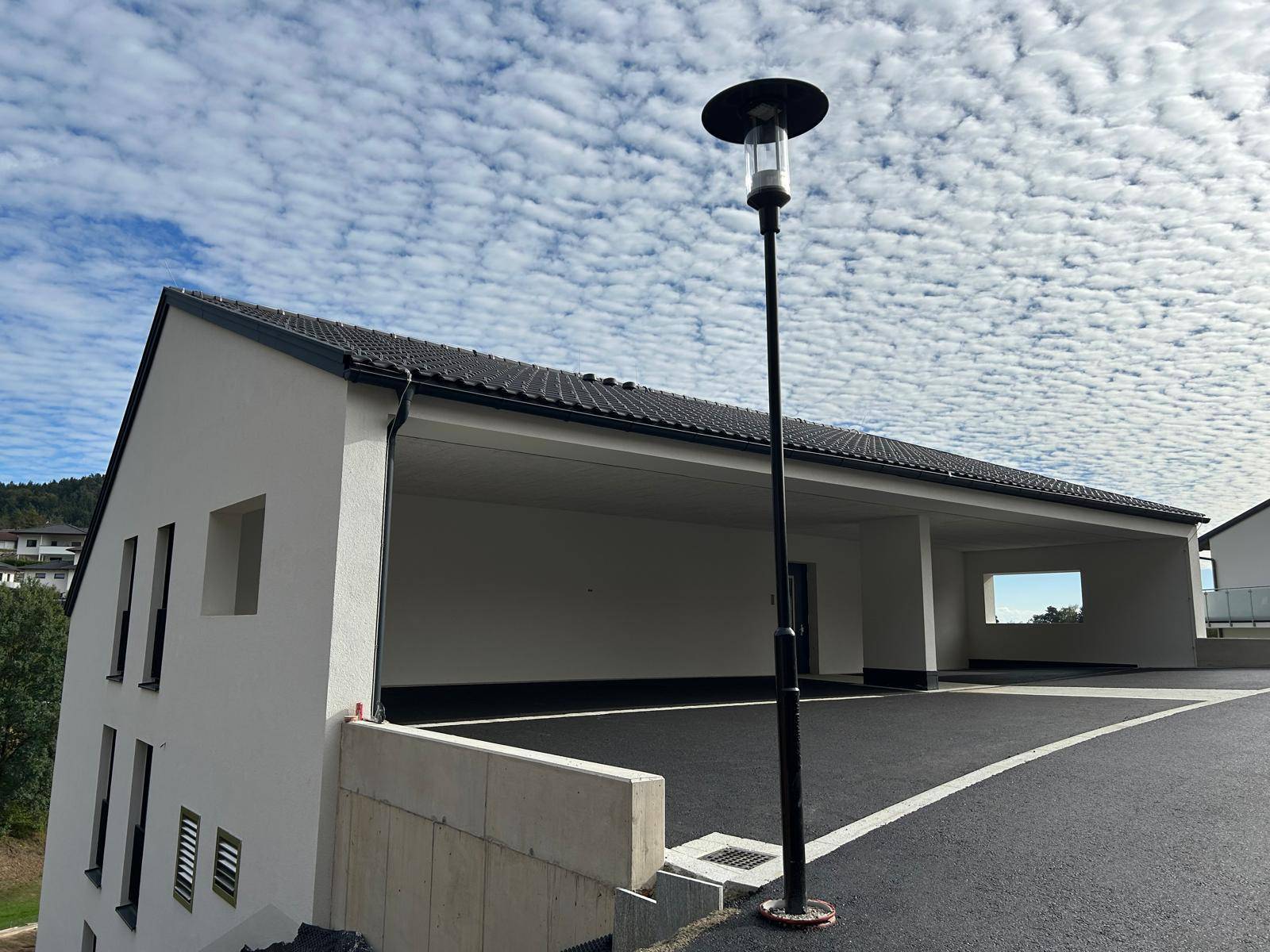 Ansicht des Neubaus mit Carport und moderner Außenbeleuchtung unter bewölktem Himmel.
