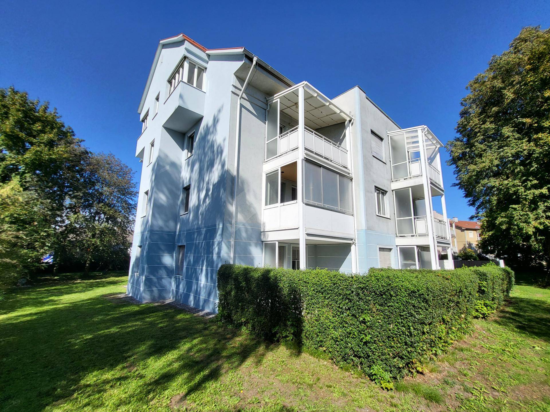 Mehrfamilienhaus mit hellblauer Fassade, mehreren Balkonen und gepflegter Grünanlage unter blauem Himmel.