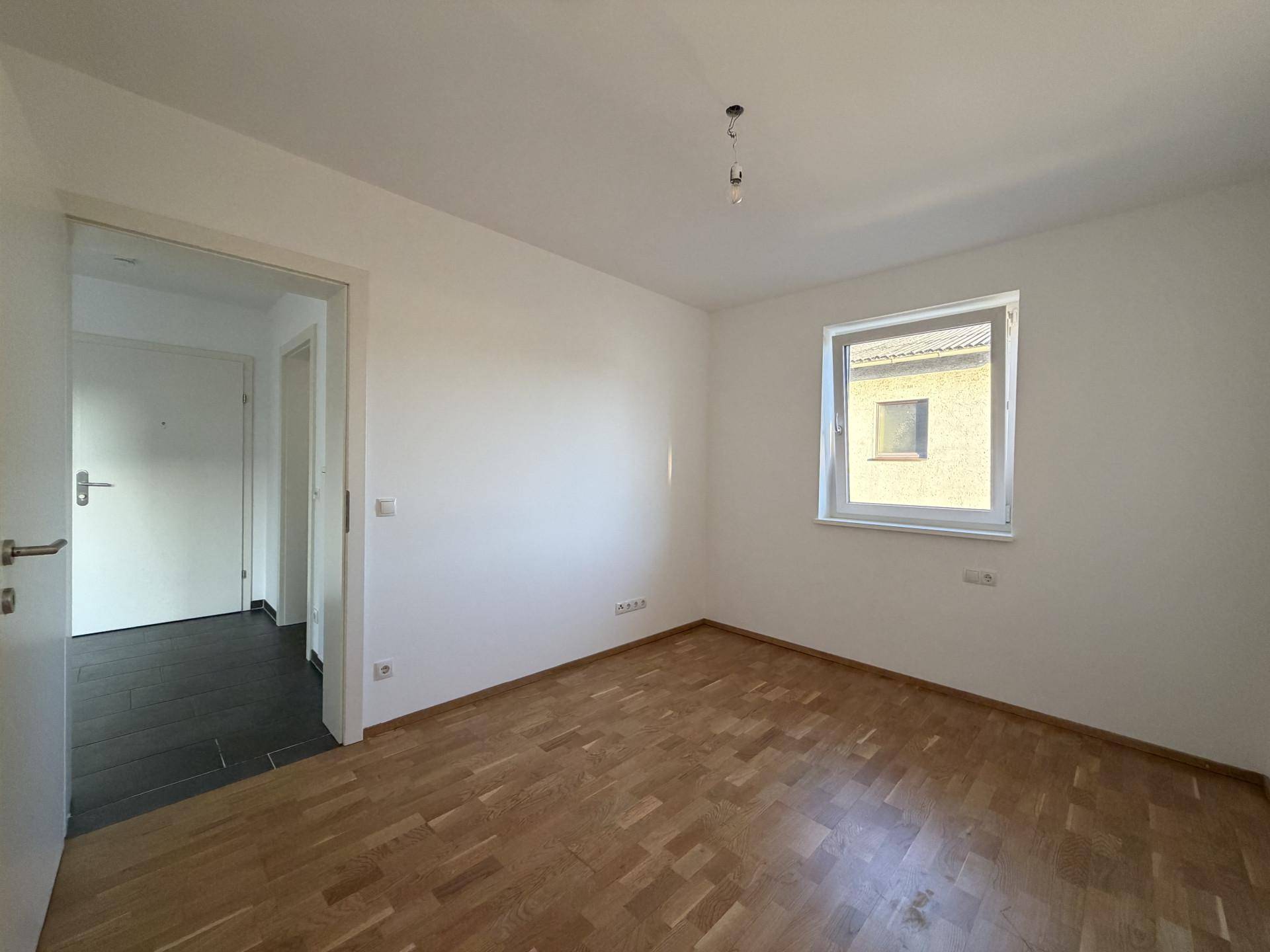 Helles Schlafzimmer mit Parkettboden und einem Fenster, angrenzend an einen gefliesten Flurbereich.