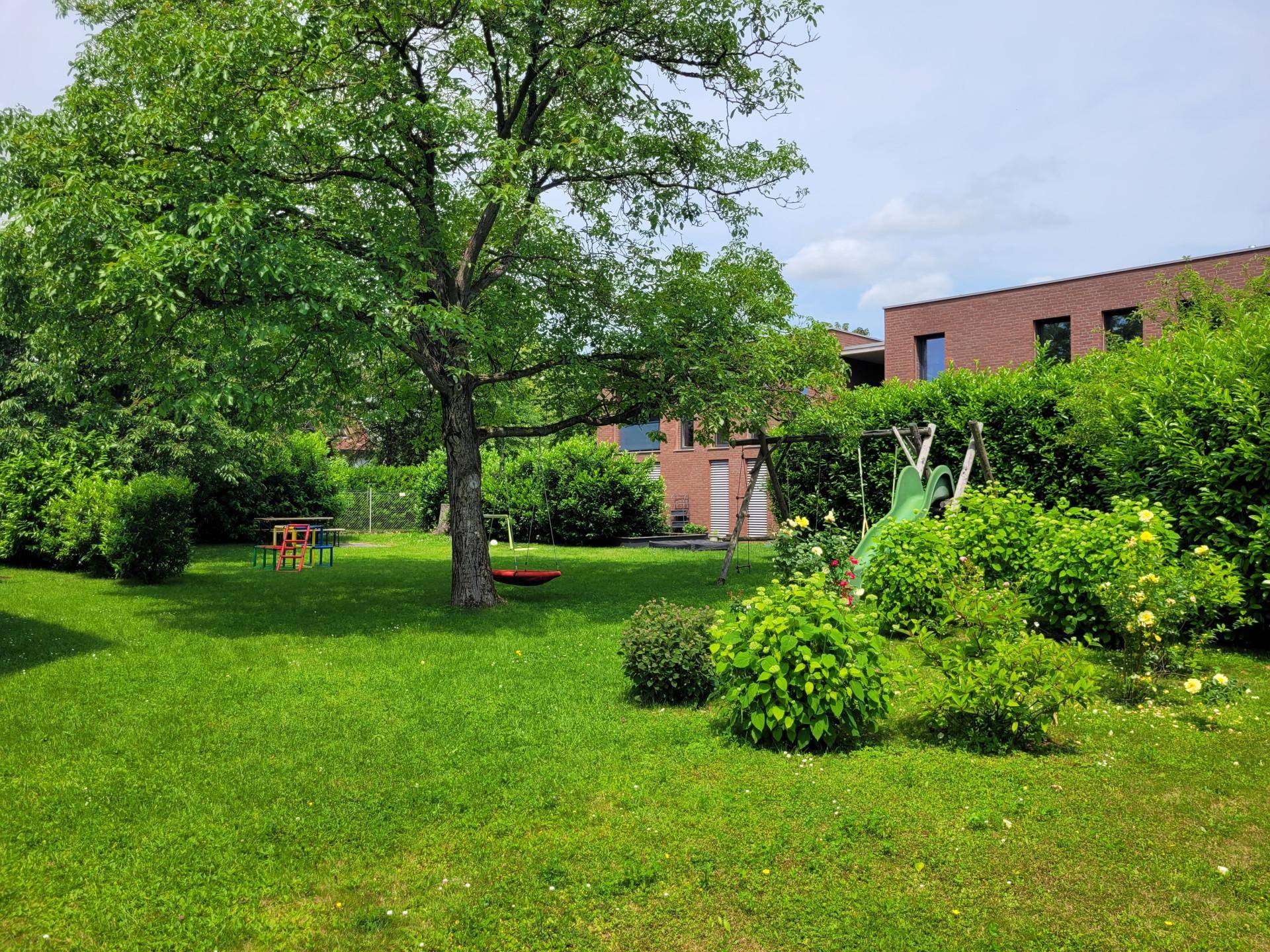 Großer, gepflegter Garten mit Rasenfläche, altem Baum, Hecken und einem Spielplatz für Kinder.