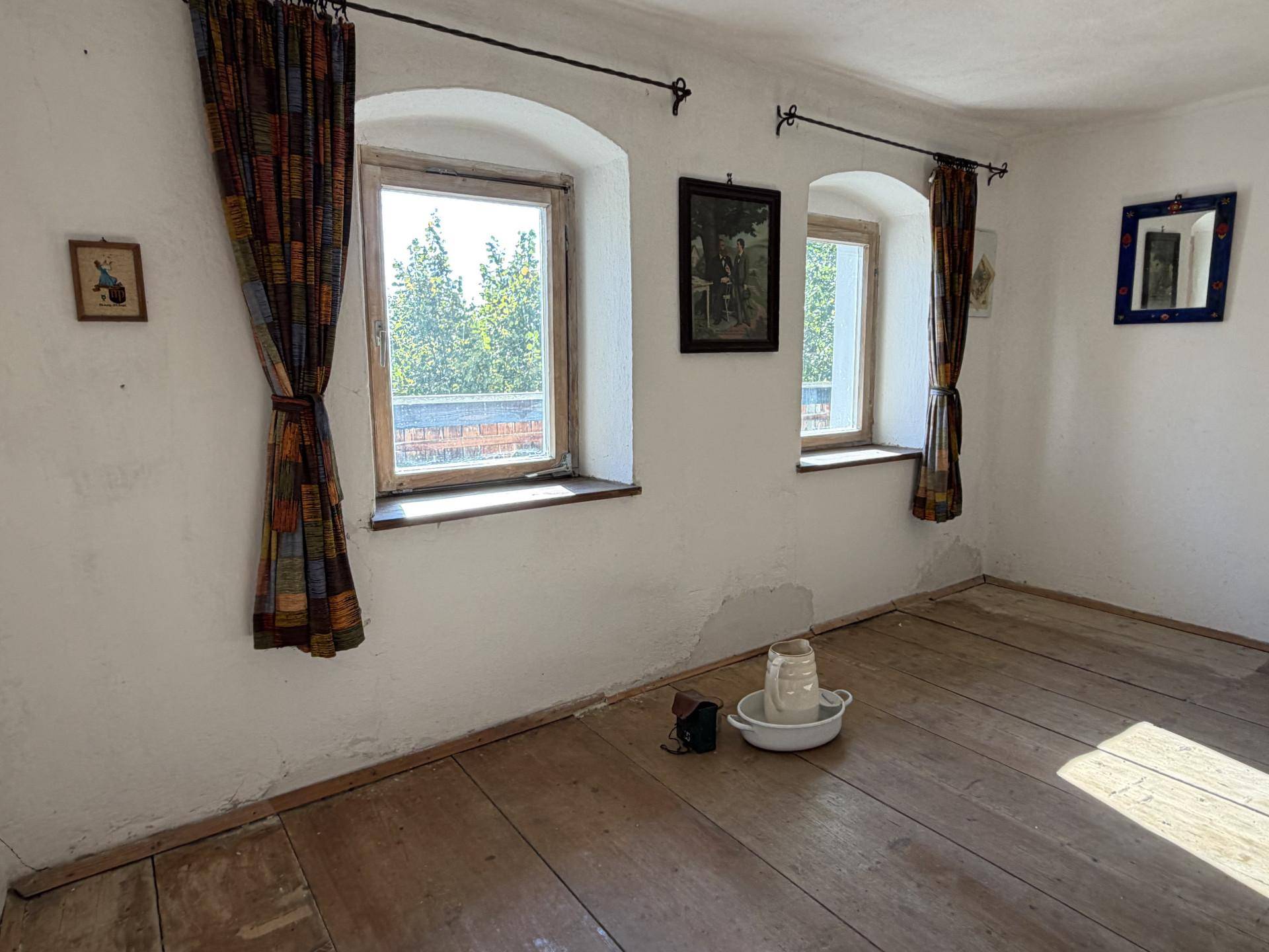 Helles Zimmer mit Holzdielenboden, zwei Fenstern und bunten Vorhängen, bereit zur Einrichtung.