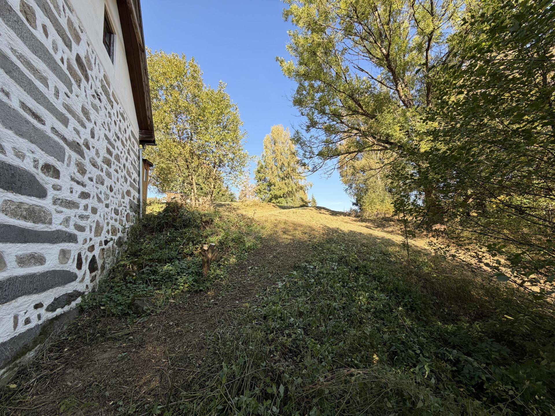 Seitenansicht eines Gebäudes mit Steinmauer und einem bewachsenen Hang unter blauem Himmel.