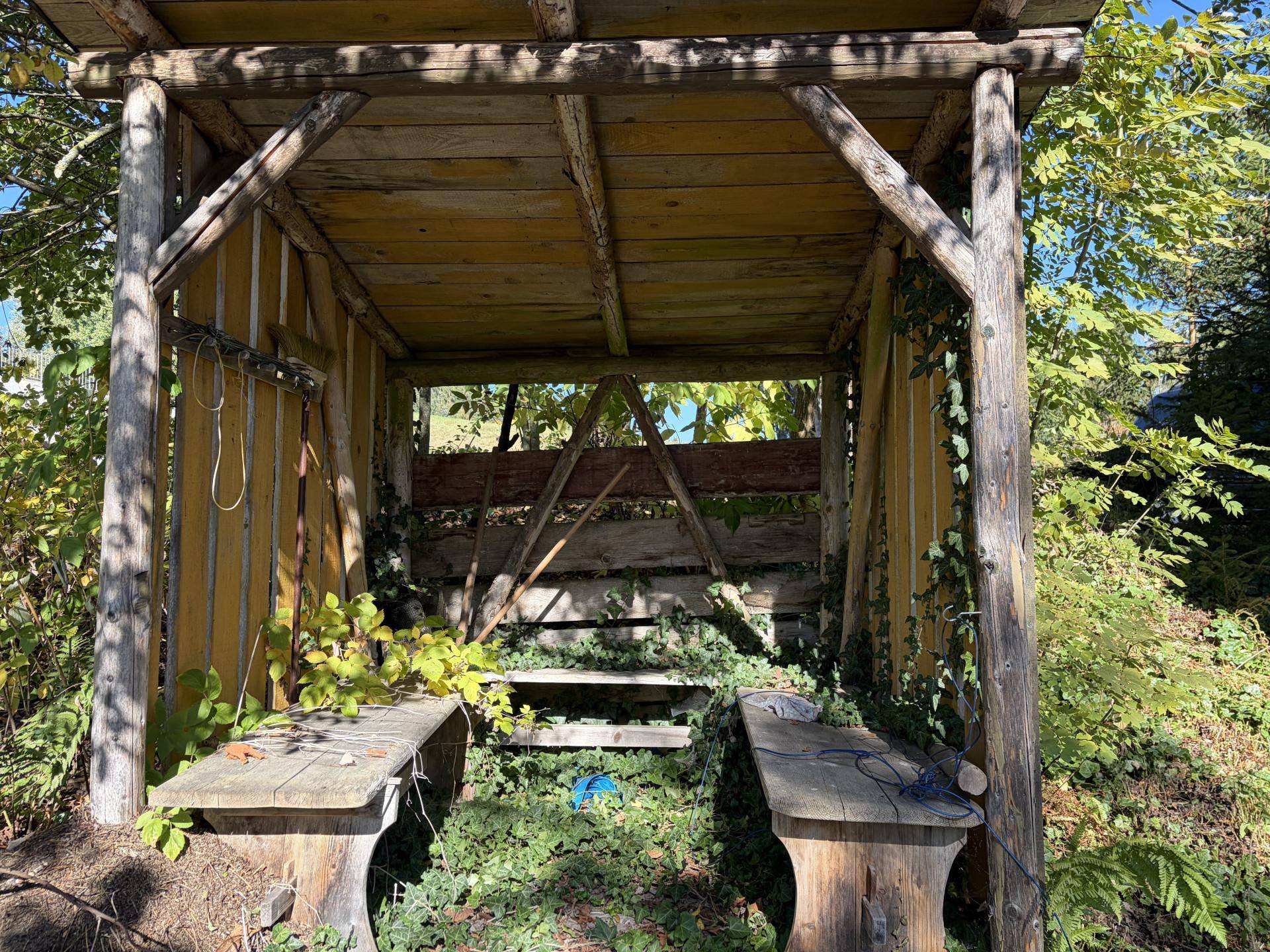 Rustikale Holzüberdachung mit Sitzbänken in einem grünen Gartenbereich.