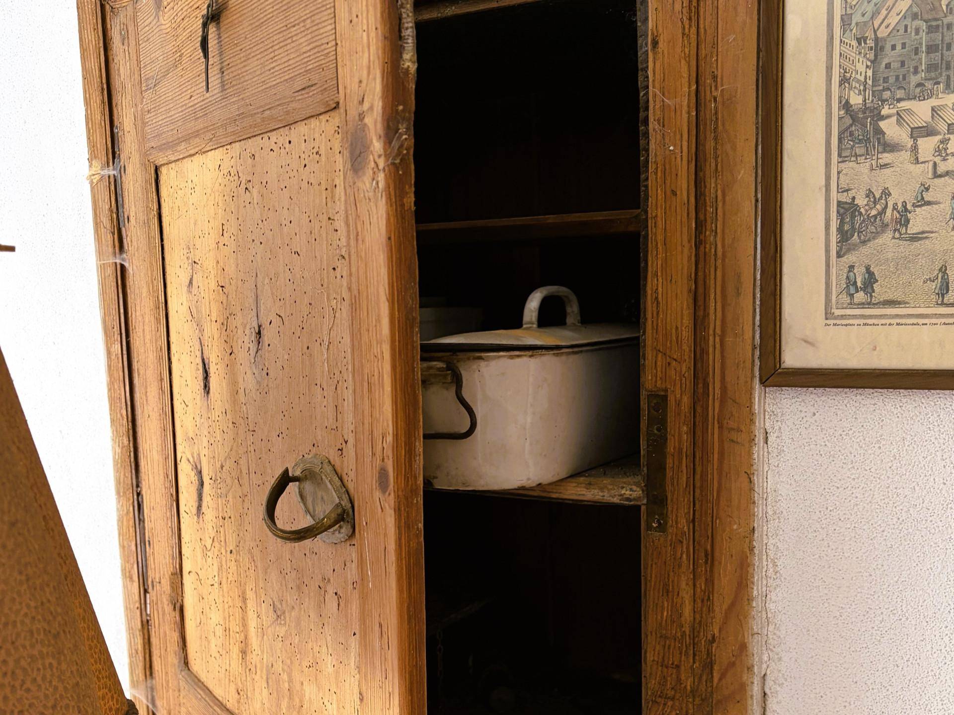 Alter Holzschrank mit geöffneter Tür, der ein weißes Kochgeschirr und ein gerahmtes Bild zeigt.