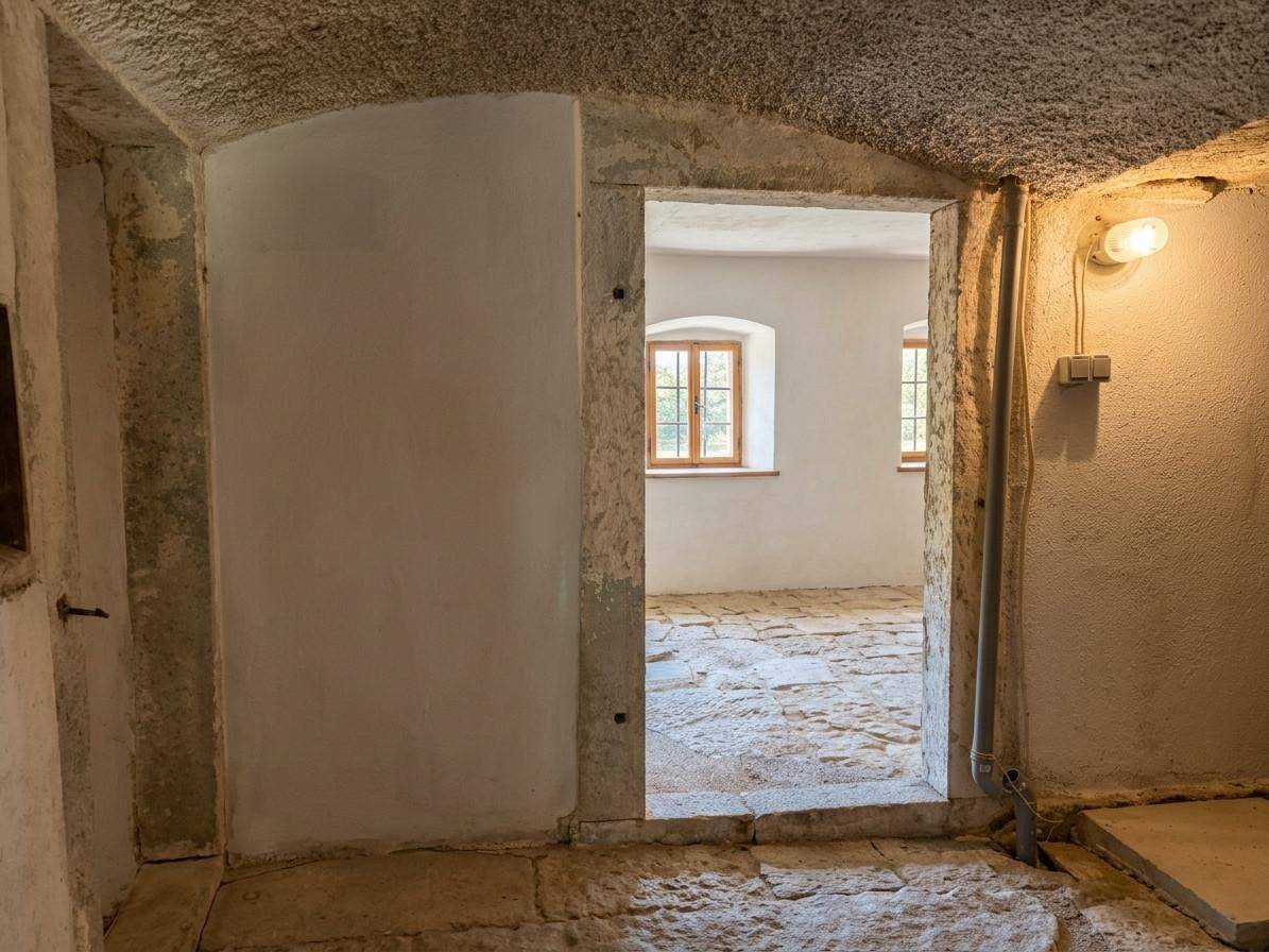 Raum mit unregelmäßigem Steinboden und einem Durchgang zu einem helleren Bereich mit Fenstern.