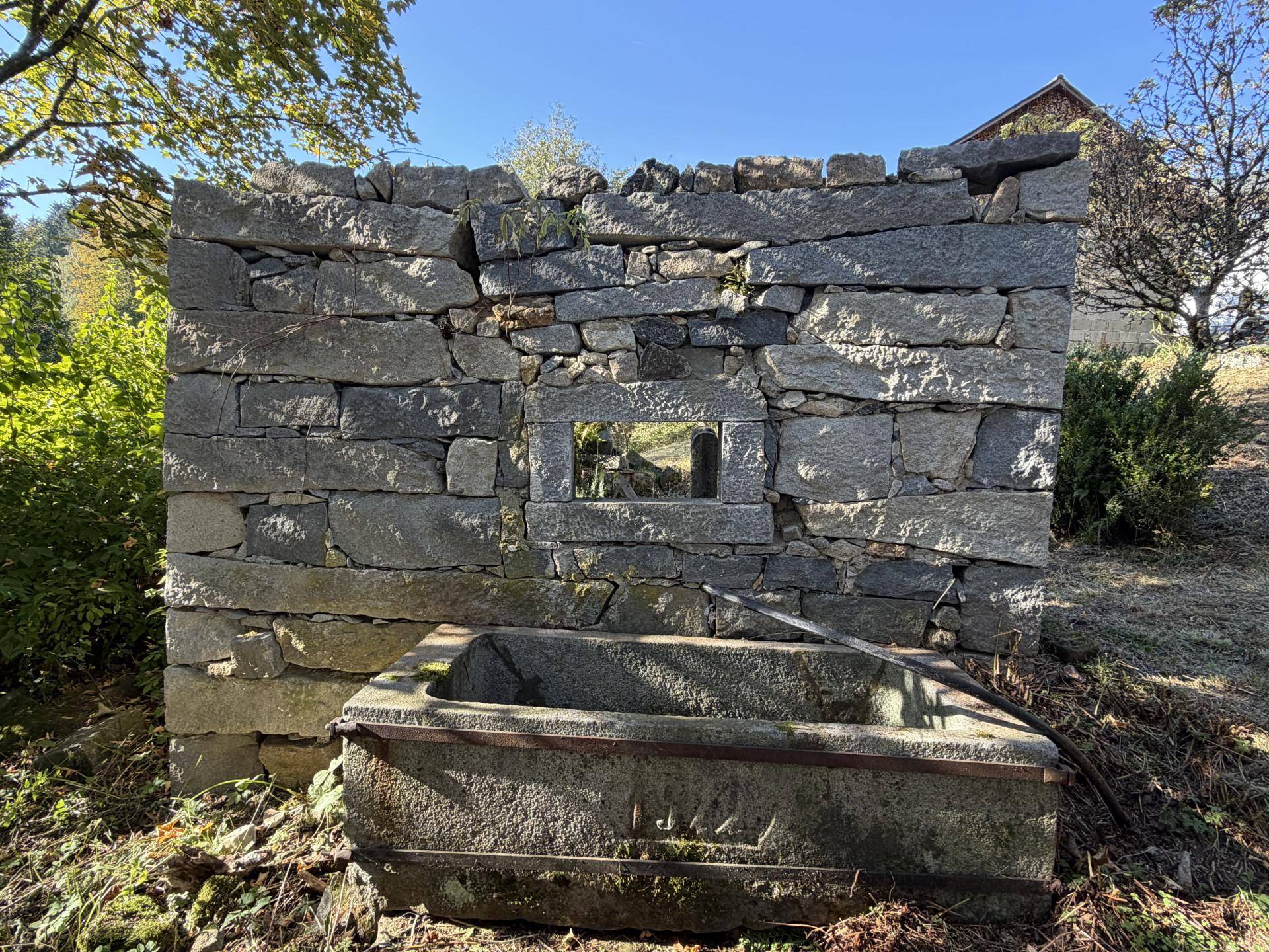 Alte Steinmauer mit integriertem Trog, umgeben von trockener Vegetation unter blauem Himmel.