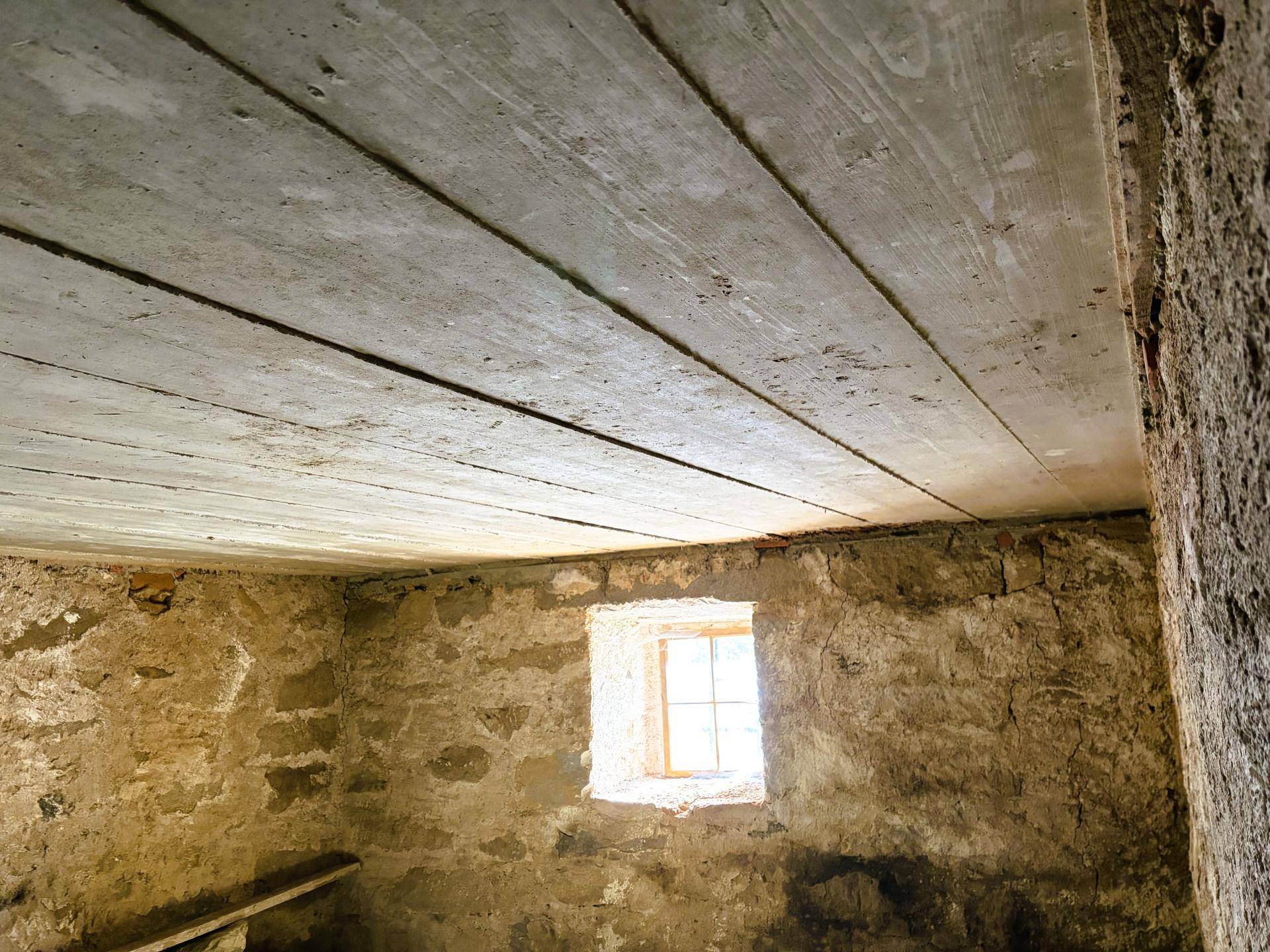 Kellerraum mit Holzbalkendecke und kleinen Fenstern, raue Steinwände.