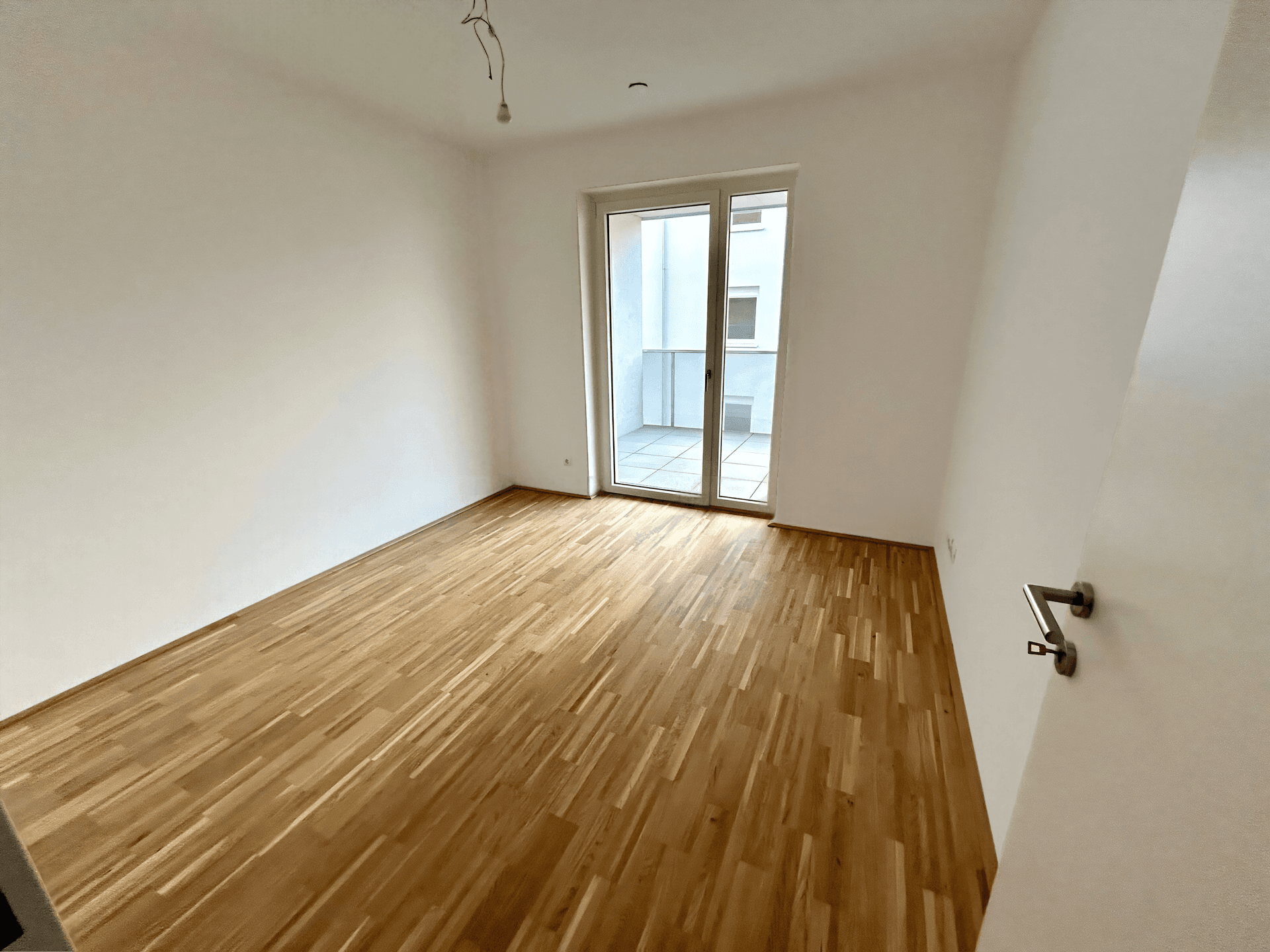 Helles, leeres Zimmer mit neuem Parkettboden und direktem Zugang zu einem Balkon.