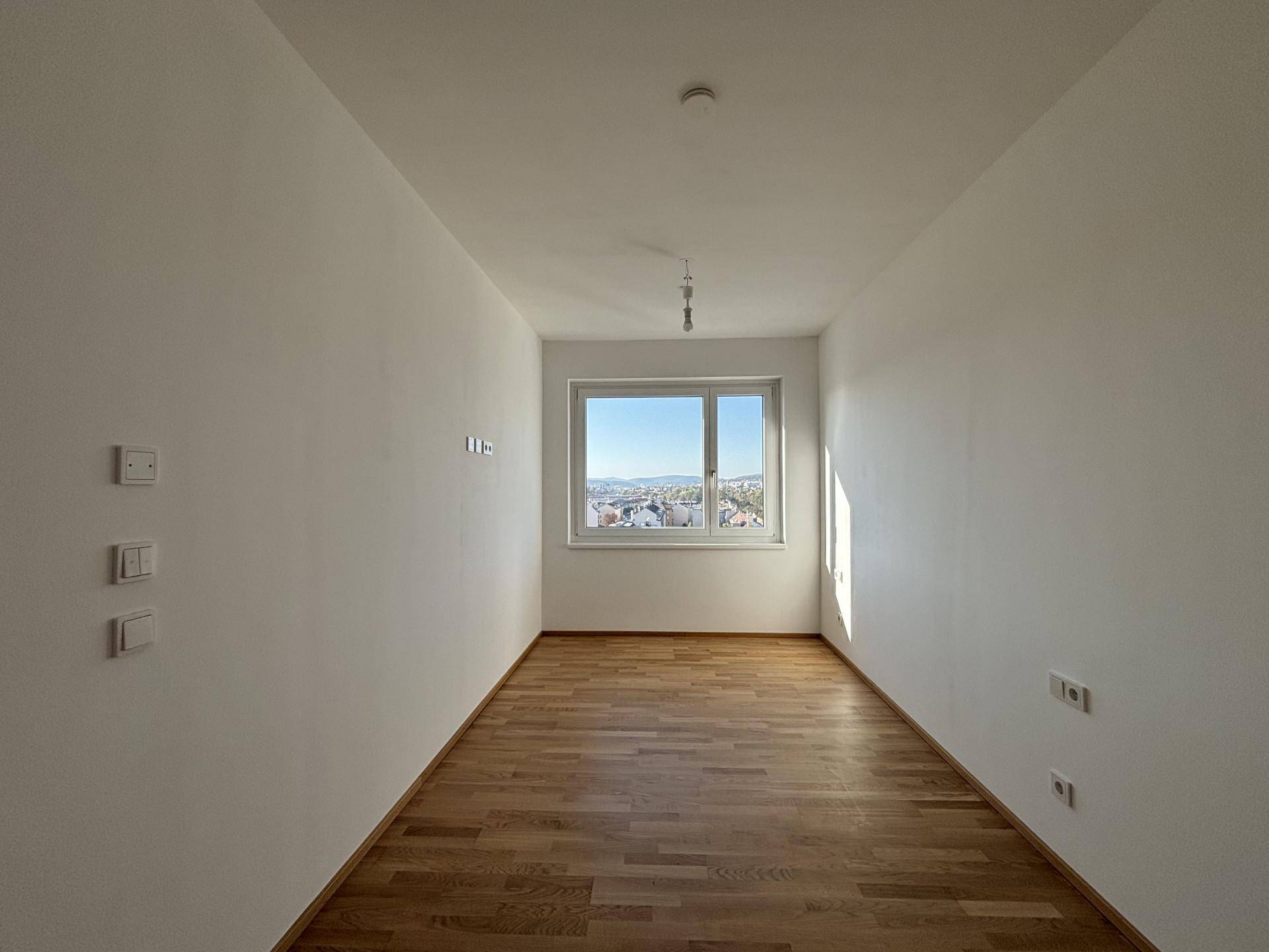Helles, leeres Zimmer mit Holzboden und einem Fenster, das einen klaren Blick auf die Stadt bietet.
