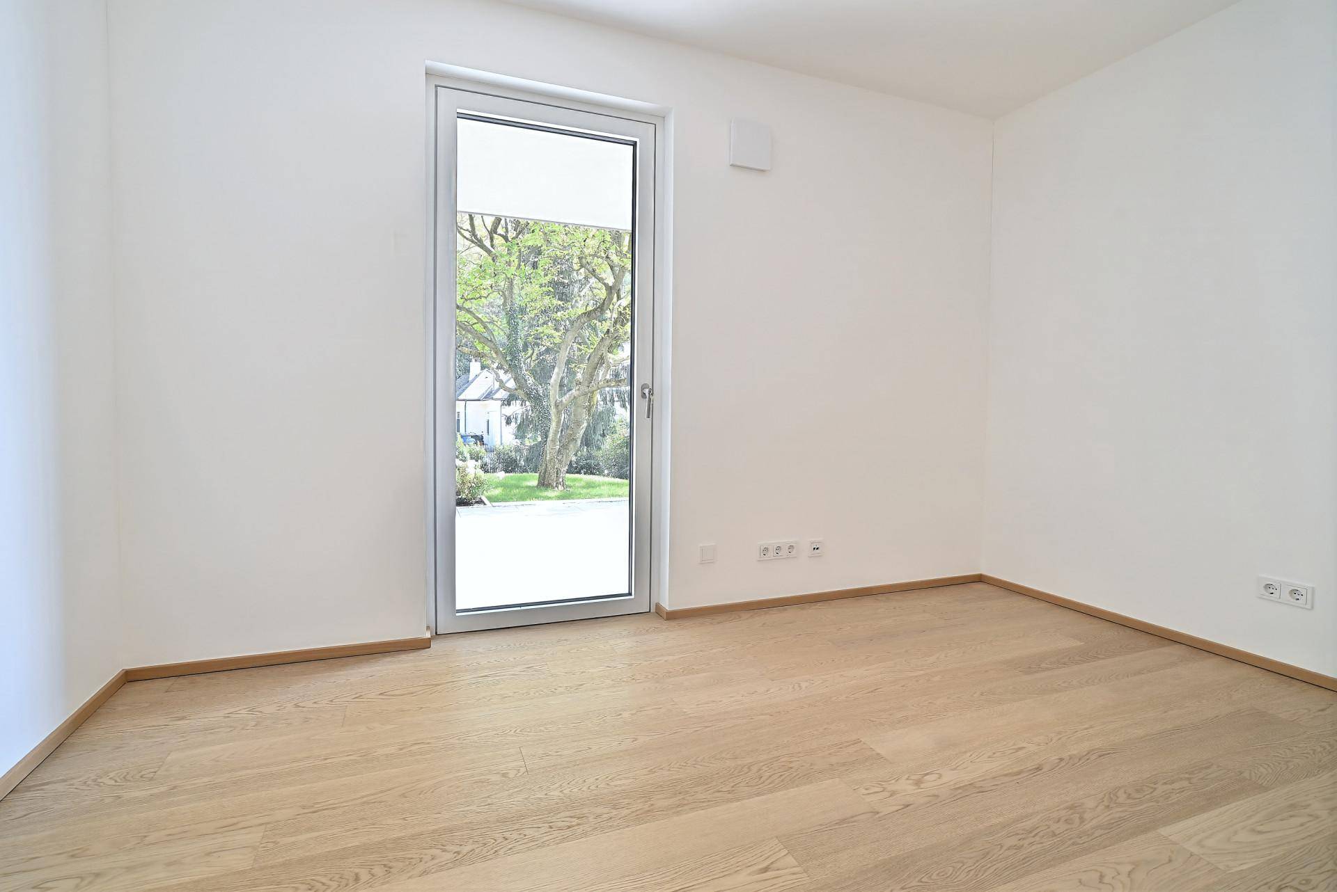 Leeres Schlafzimmer mit Holzboden und großer Fenstertür, die viel Tageslicht hereinlässt und zum Garten führt.