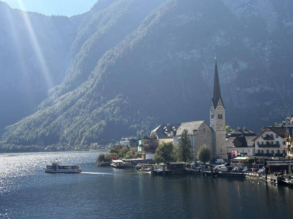 Malerisches Dorf am See mit Kirchturm und Bergen im Hintergrund, ein Boot fährt auf dem Wasser.