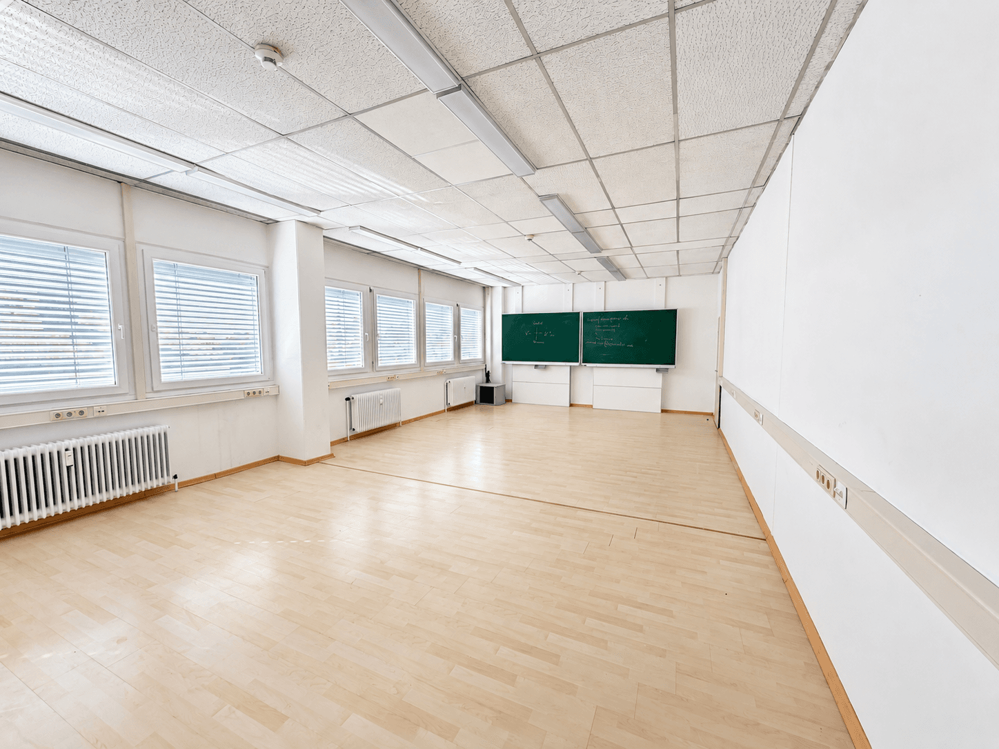 Geräumiger, leerer Klassenraum mit hellen Böden, großen Fenstern und grünen Tafeln an der Wand.